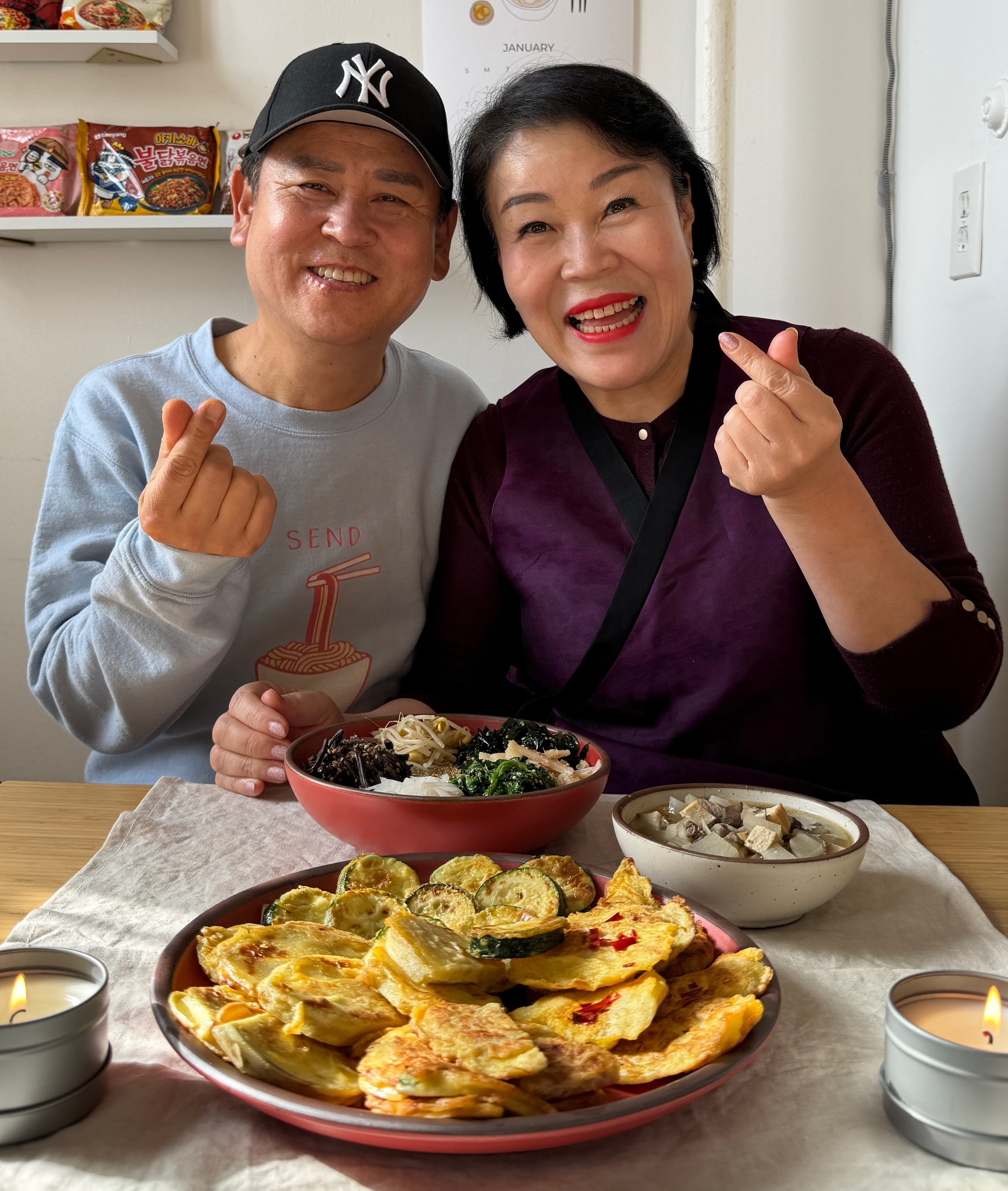 A mom and dad are smiling at the camera behind a table filled with Korean Lunar New Year foods plated on ceramic plates and bowls.