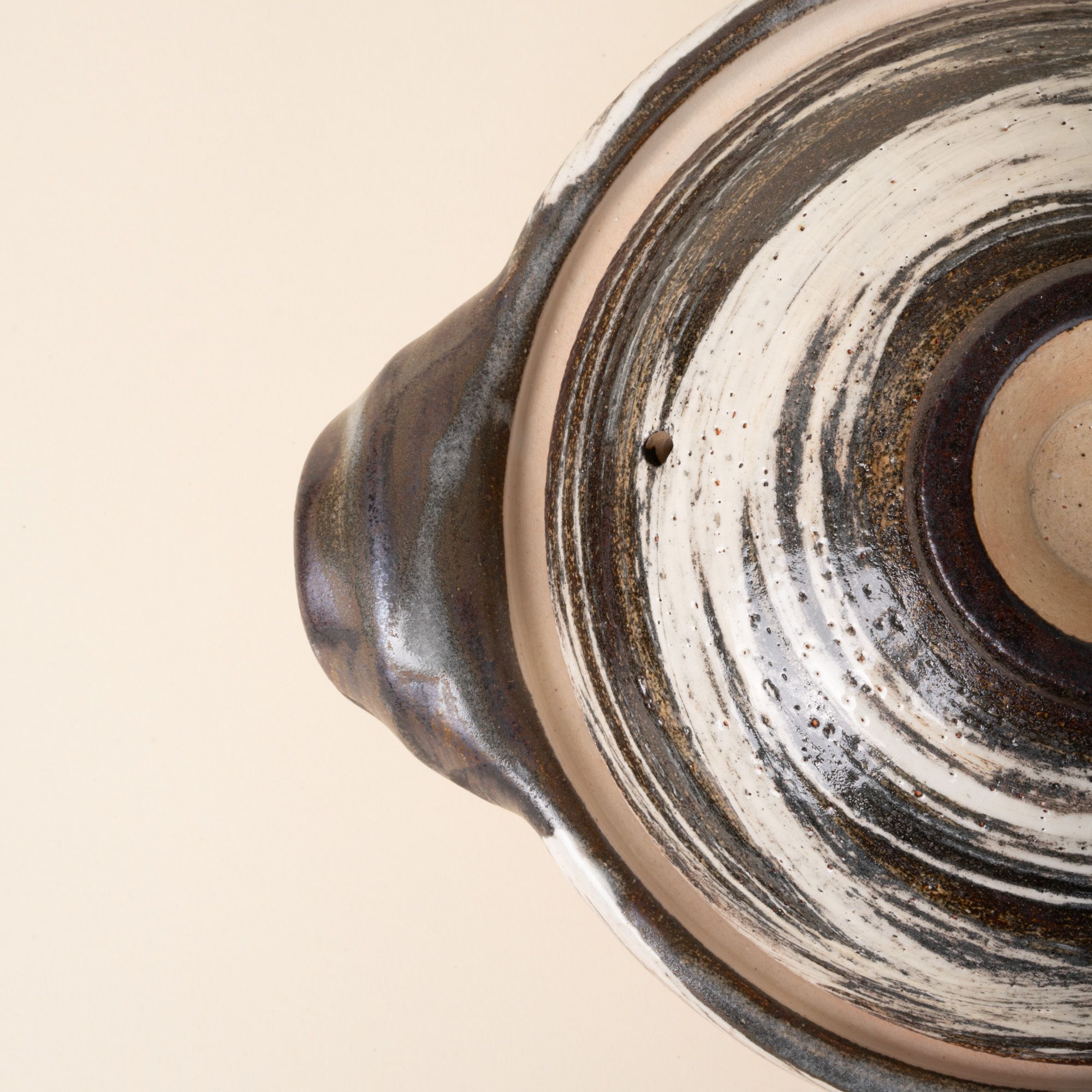 An overhead closeup view of a ceramic donabe with side handles, finished in a black-and-white brushed glaze over natural clay.