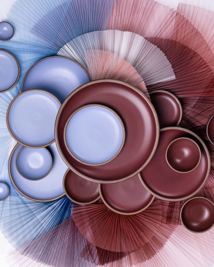 Ceramic plates and bowls in periwinkle and plum colors are artfully arranged against pleated tulle fabric