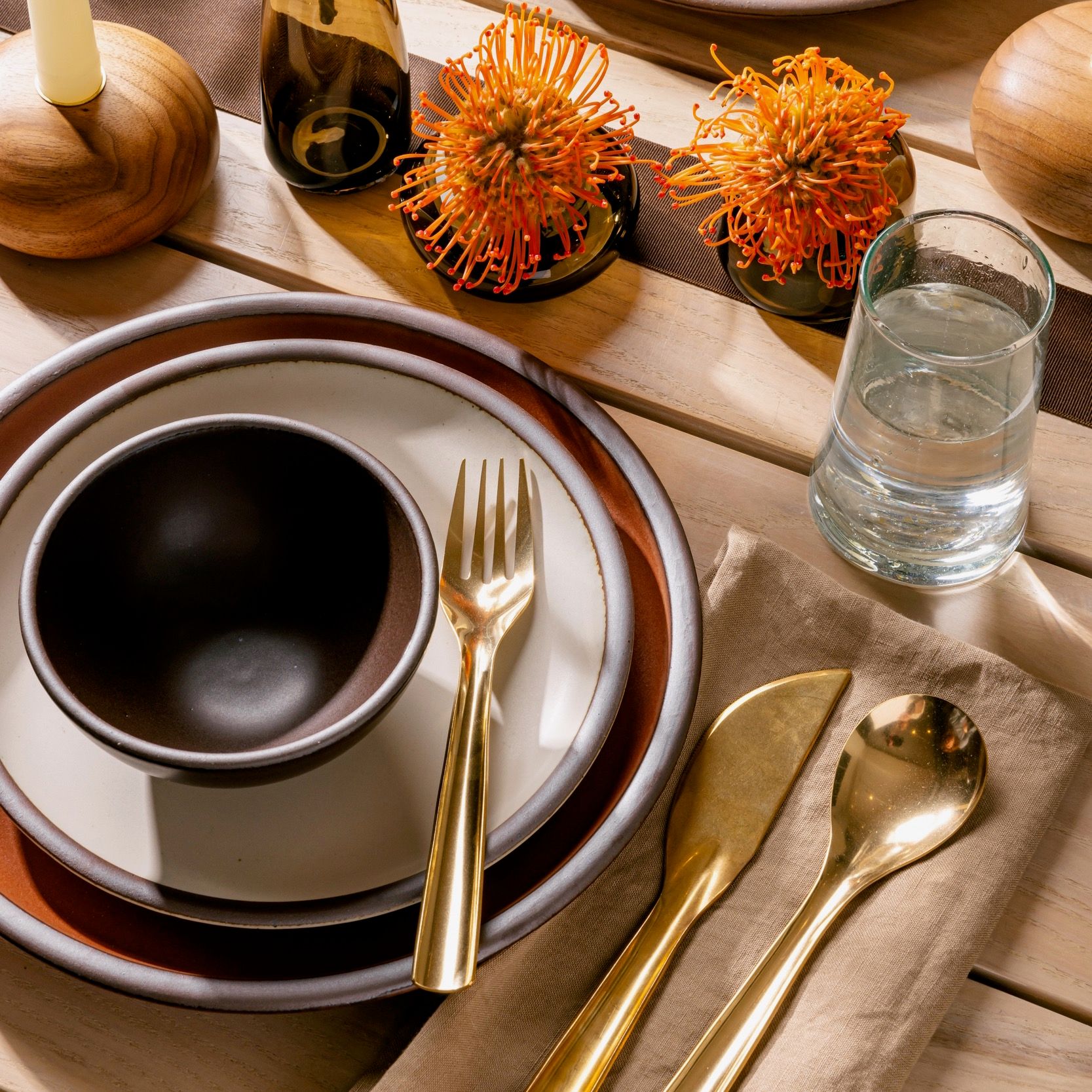 An overhead view of a table setting with ceramic bowls and plates, brass flatware, a water glass, and a dried floral arrangement in amber glass vases.
