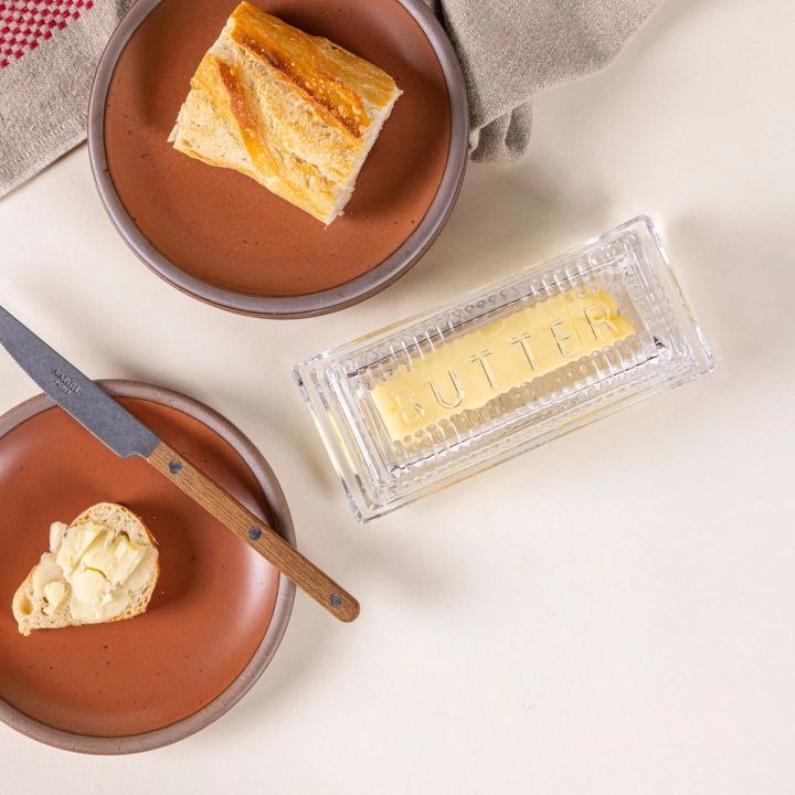 Traditional glass butter dish with 'Butter' on top and butter inside. Off to the side are terracotta plates with bread and a knife.