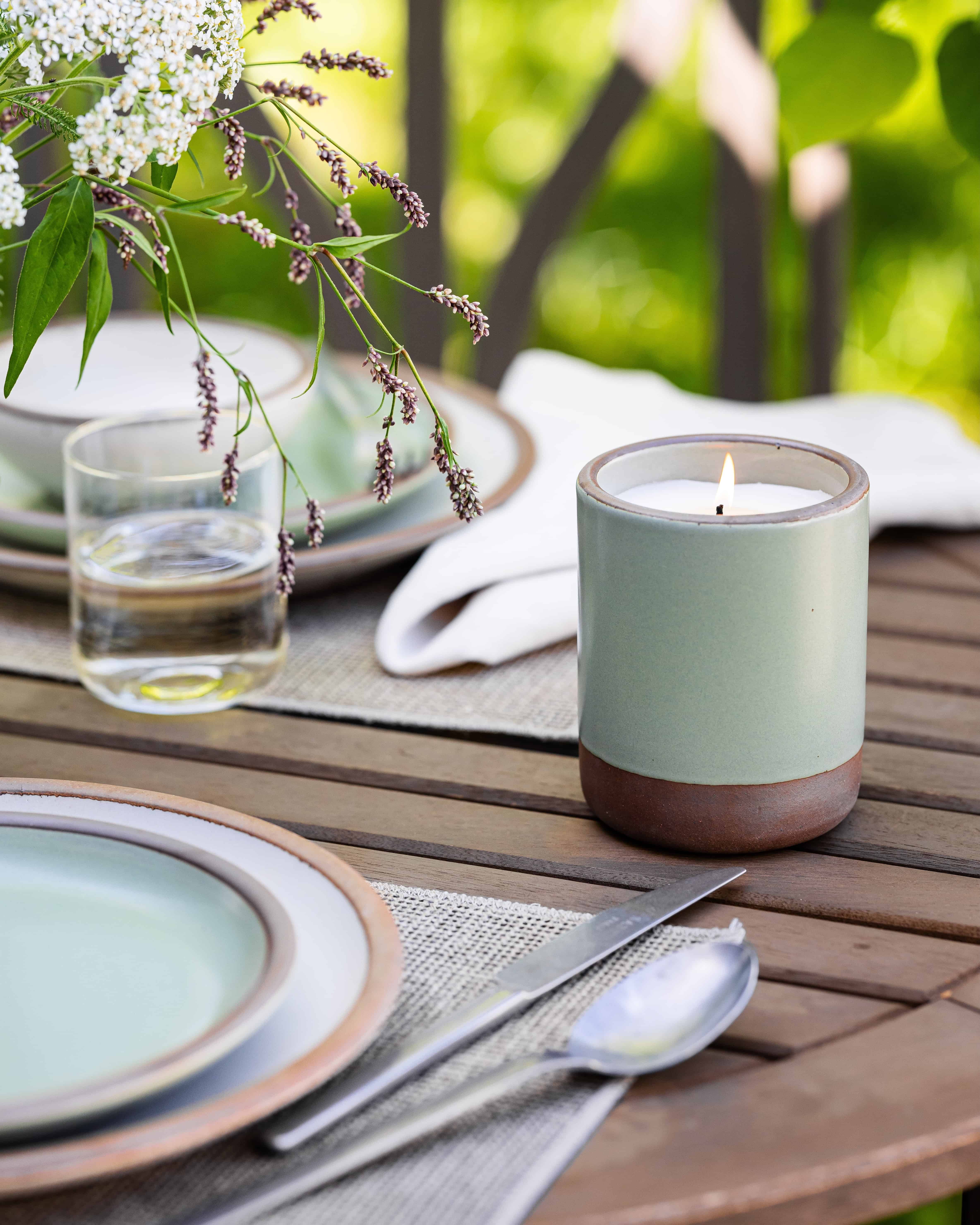 On an outdoor table, there is a few place settings and an outdoor candle in a ceramic cylindrical vessel in a sage green color that is lit.
