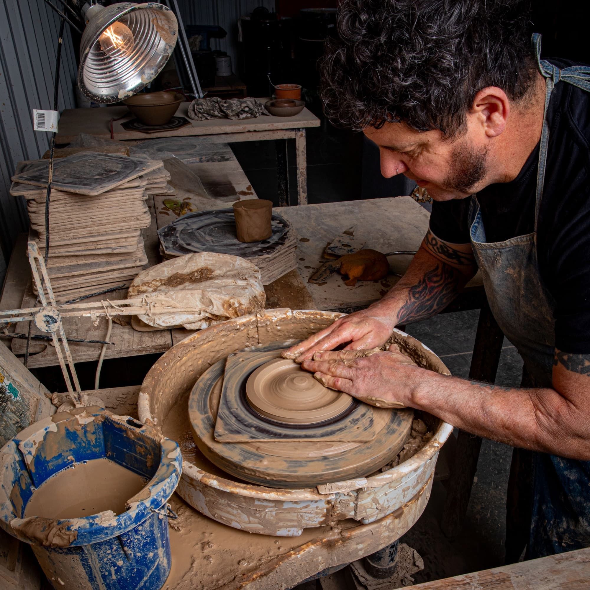 In a pottery workshop, a person sits at a potter's wheel shaping a plate-shaped incense holder 