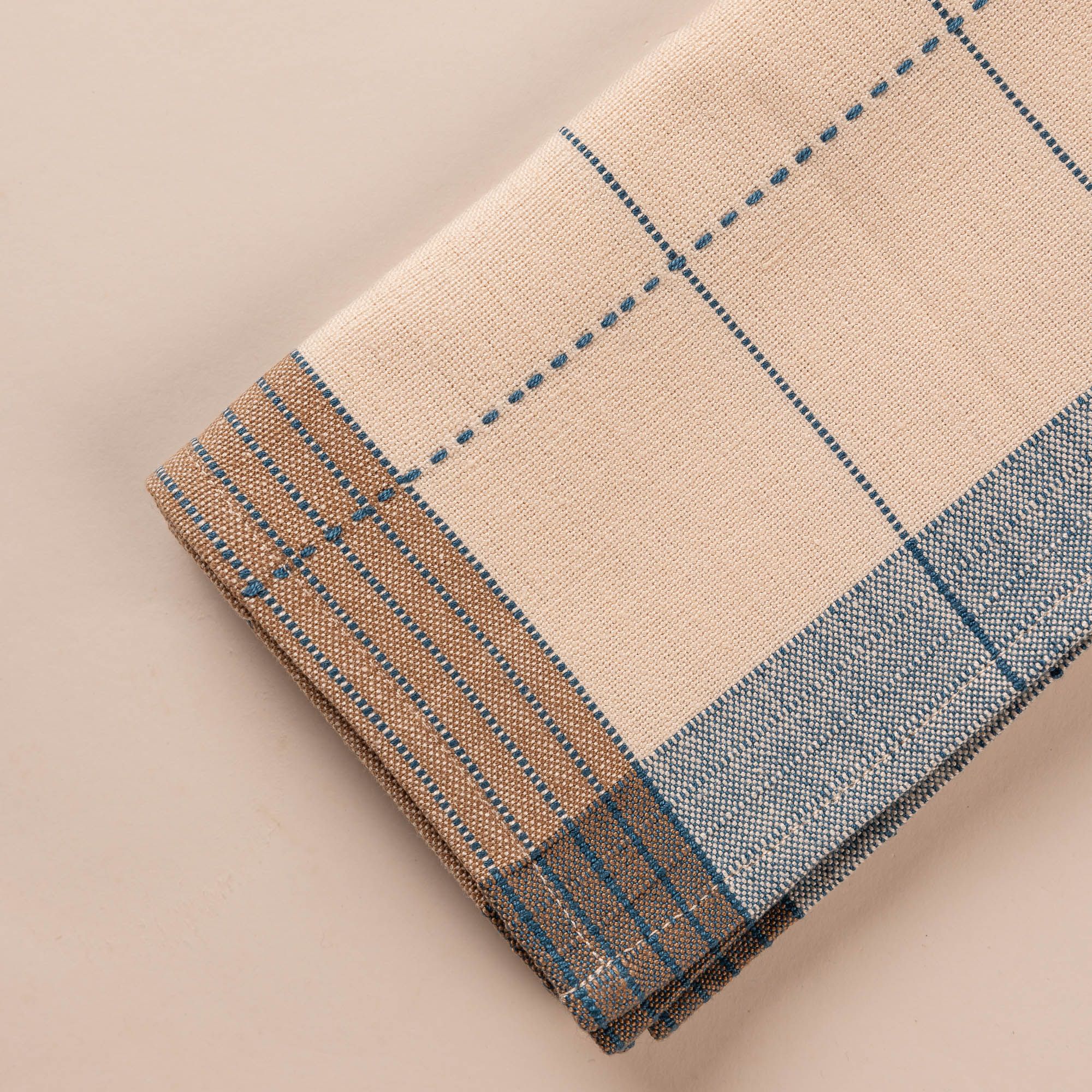 A closeup of a natural folded napkin designed with turquoise gridlines with light blue and tan striped edging.