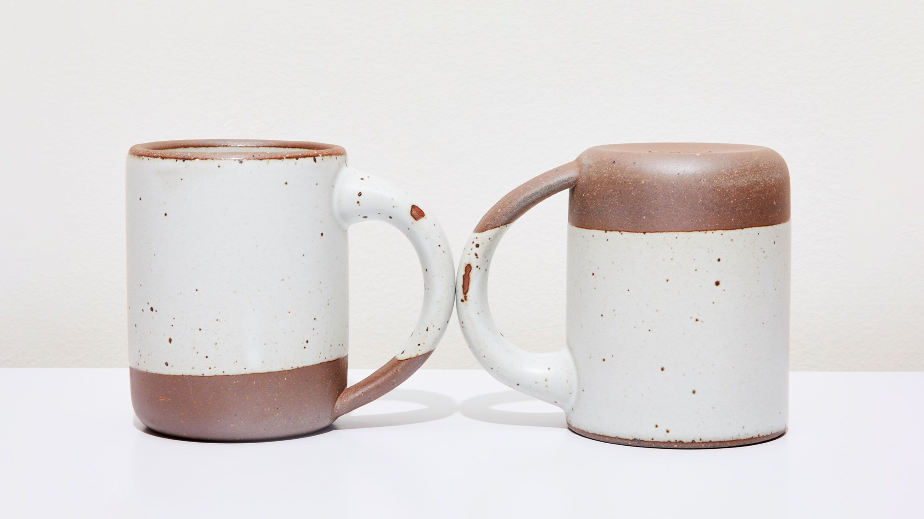 Two white ceramic mugs, one sitting up right, one upside down, with little glaze chips on the handles.