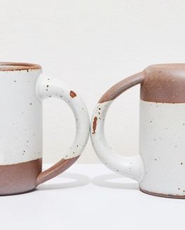 Two white ceramic mugs, one sitting up right, one upside down, with little glaze chips on the handles.