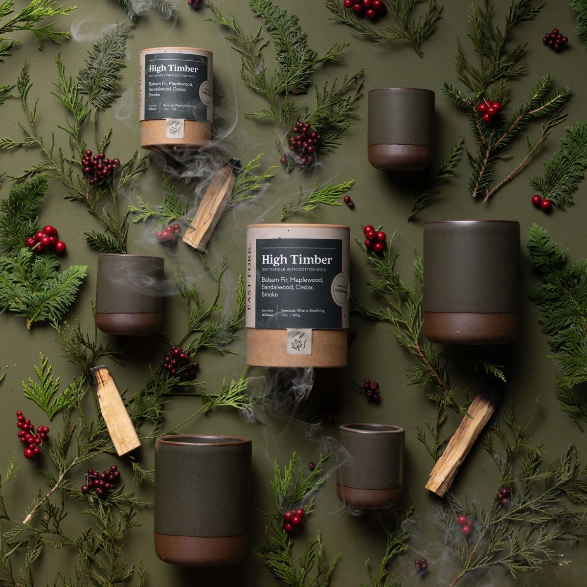 An artful layout of The Candle in High Timber - centered is a cardboard packaging tube with the candle's name, surrounded by small and large ceramic candles in a cylindrical vessel in a pine green color, surrounded by pines, berries, and cedar