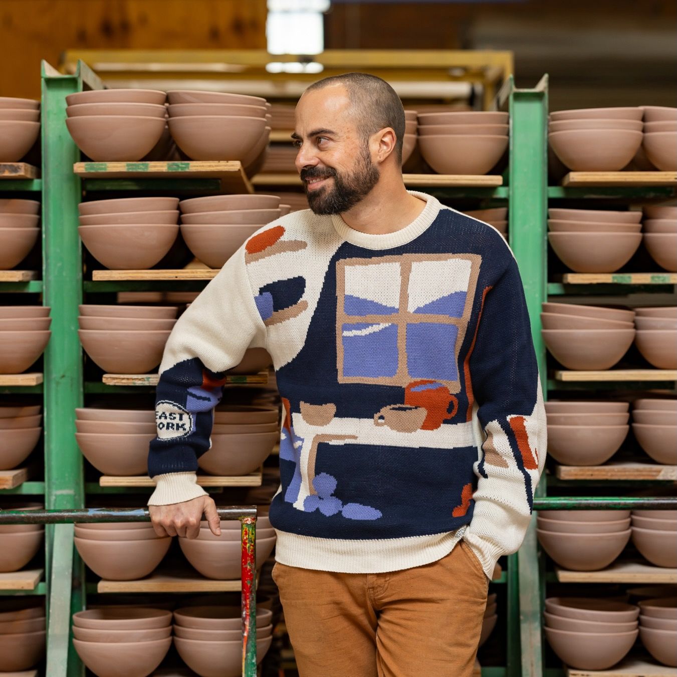 Alex wears a sweater with an illustration of a pottery workshop with a large window, shelves, and table with mugs and bowls. Alex stands in front of a rack of ceramic bowls in progress.