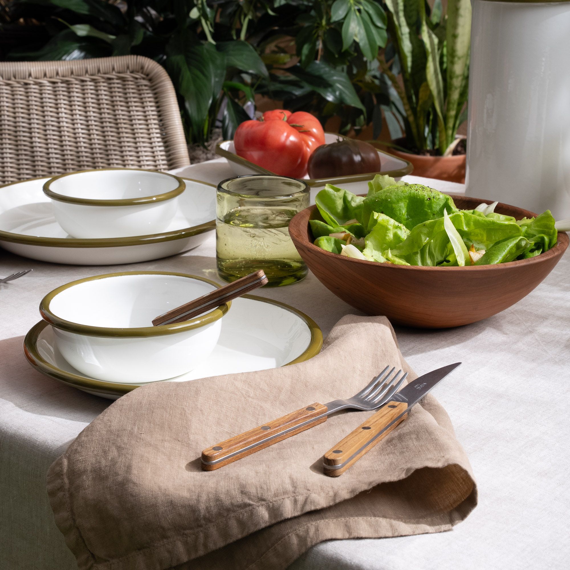 A tabletop filled with white enamel plate and bowl table setting with flatware with wood handles, along with a napkin, wood bowl, and glass.