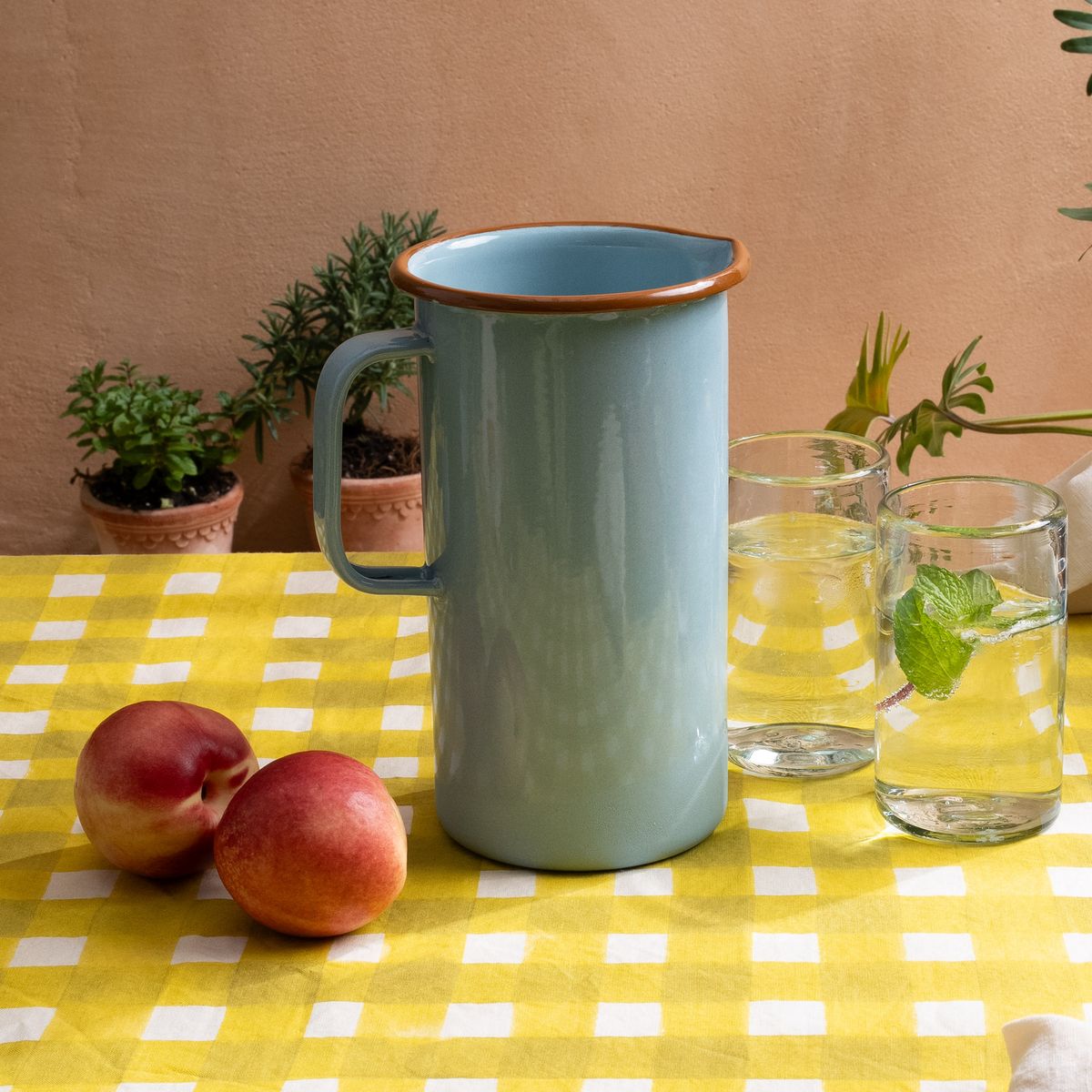A large enamel pitcher in a robin's egg blue sitting on a bright yellow crosshatch tablecloth with tall clear glasses, and apples.