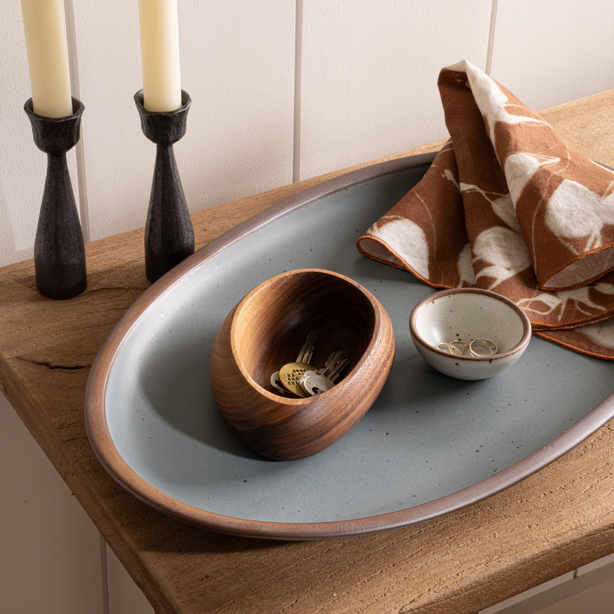 An oval ceramic tray holds a wooden bowl with keys, a small dish with rings, and a patterned cloth beside two tall black candlesticks.
