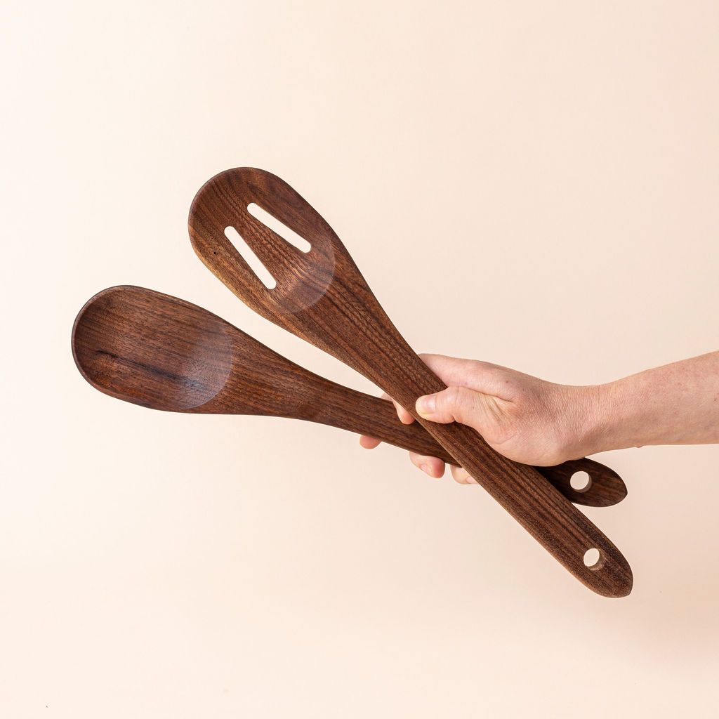 Hand holding two large wooden spoons in a deep brown color - one is solid and one has two slots, both have a hole on the end for hanging.
