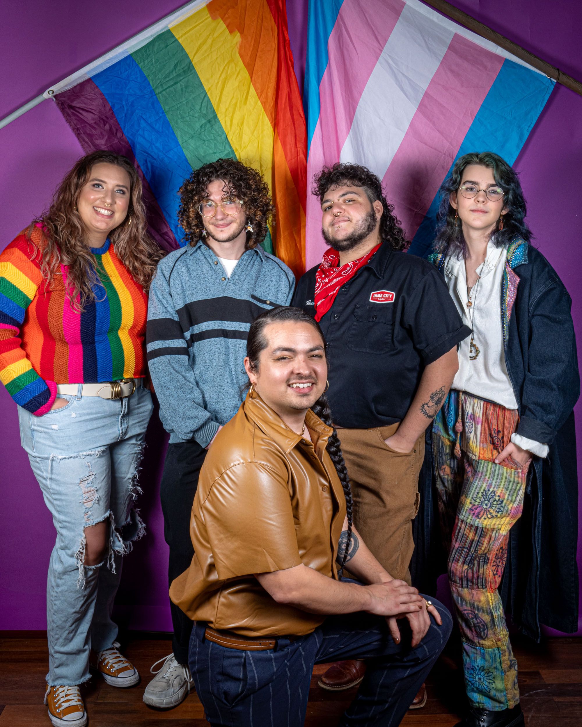 5 people are gathered together smiling in front of pride and trans flags and a purple background.