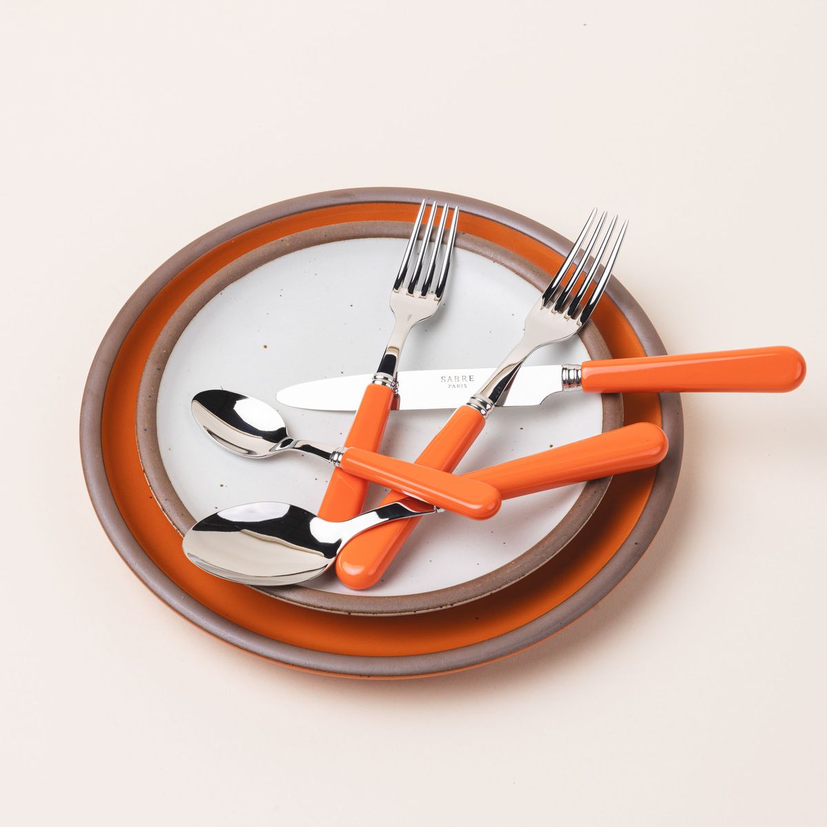A five piece flatware set with shiny utensils and matte bold orange handles sits on a bold orange and white ceramic plates