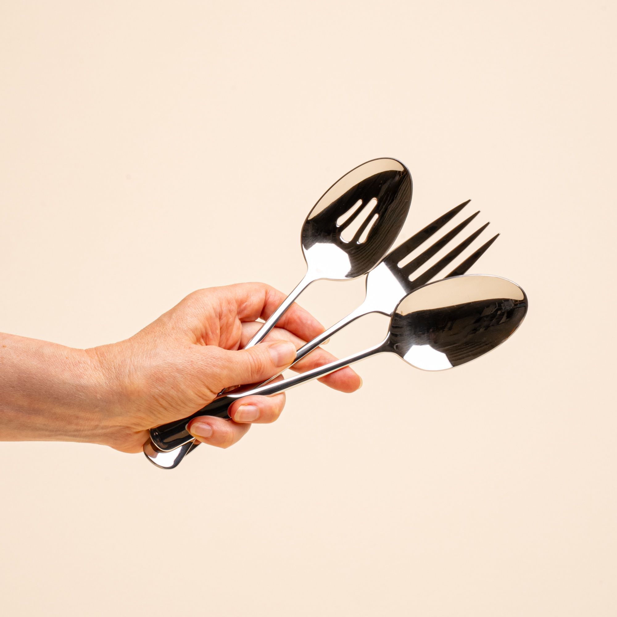 A hand holds three stainless steel serving utensils including a serving fork, serving spoon, and pierced spoon in a polished finish with a classic design