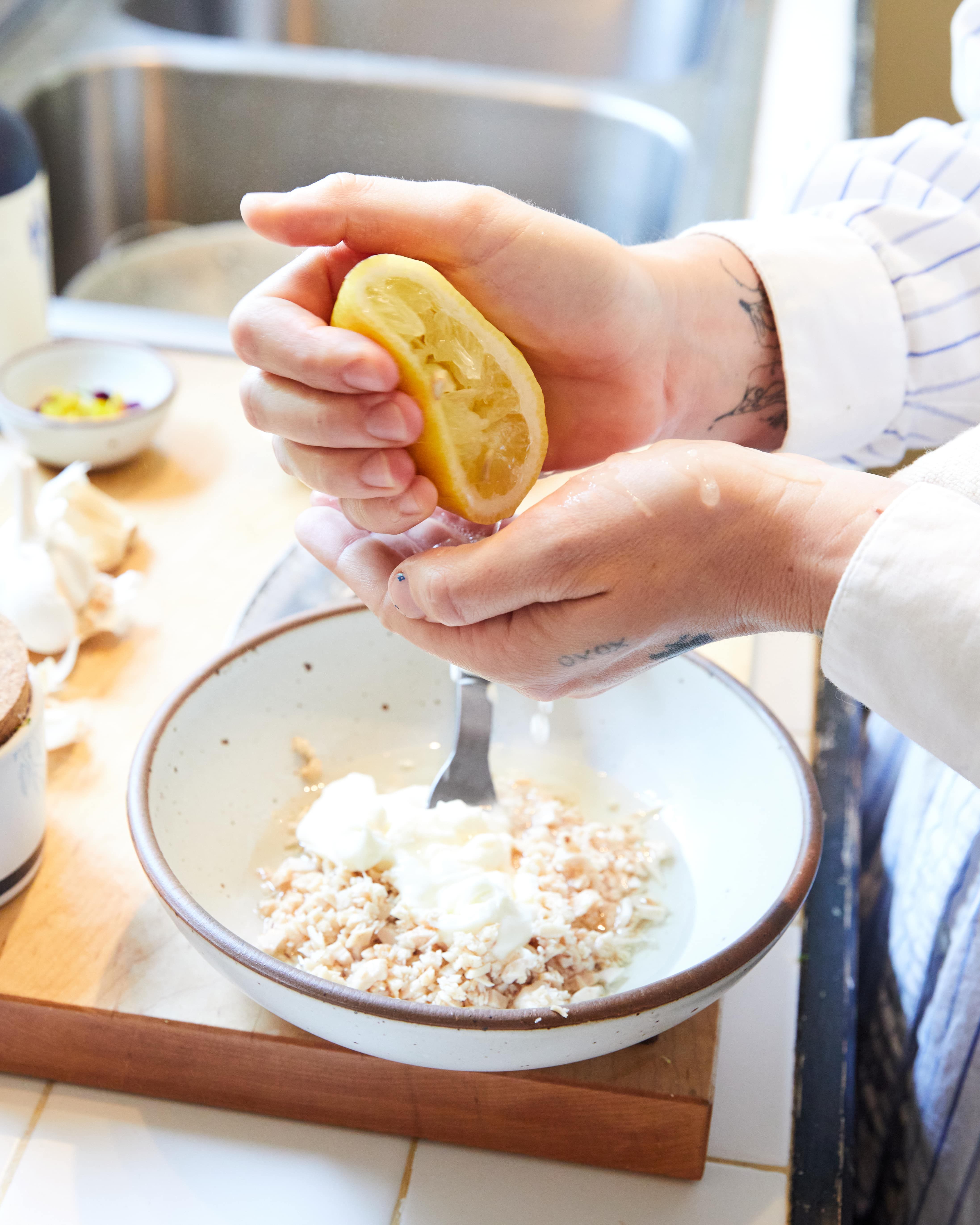 A person squeezes lemon juice out of a lemon into their hand above a bowl of ingredients