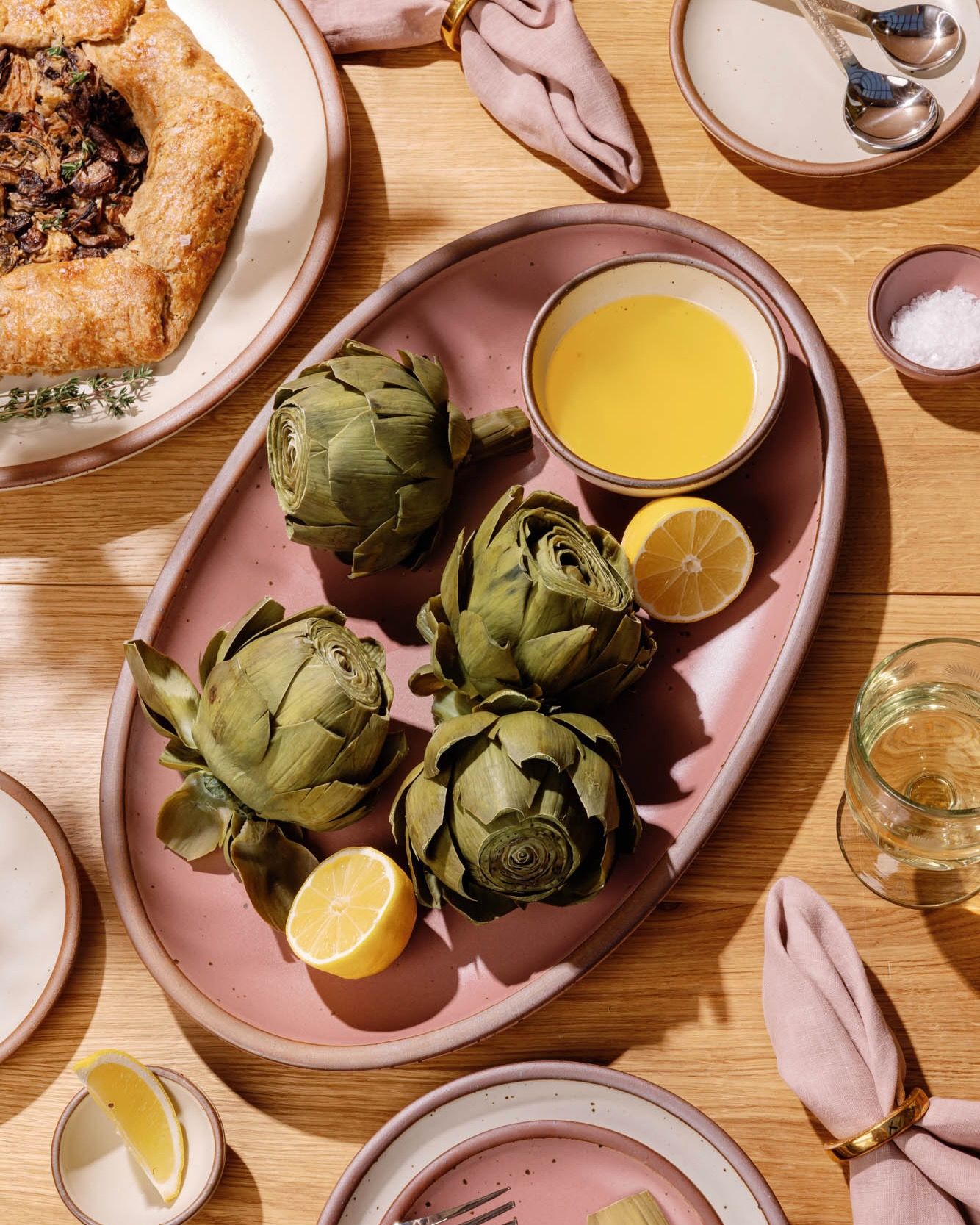 Artichokes on a pink oval platter with lemon halves and a bowl of melted butter on a wooden table.