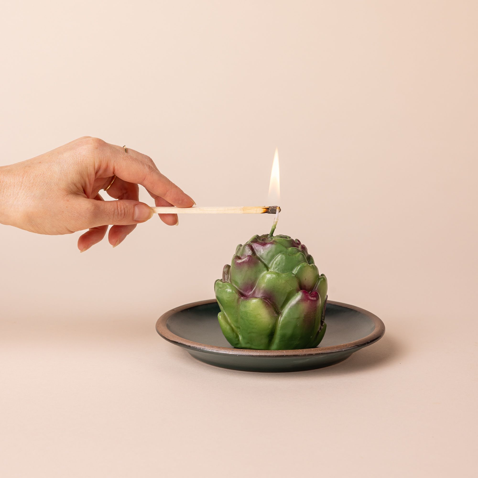 An artichoke candle with a burning flame sits on a small plate. A hand has a lit match igniting the flame.