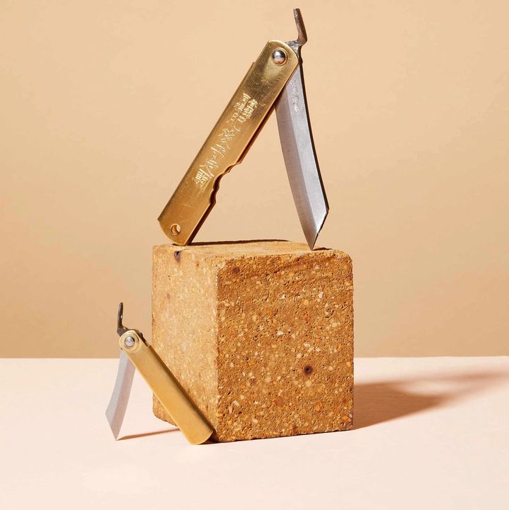 Two brass pocket knives, one small and one large, balancing on edges, with a cork block