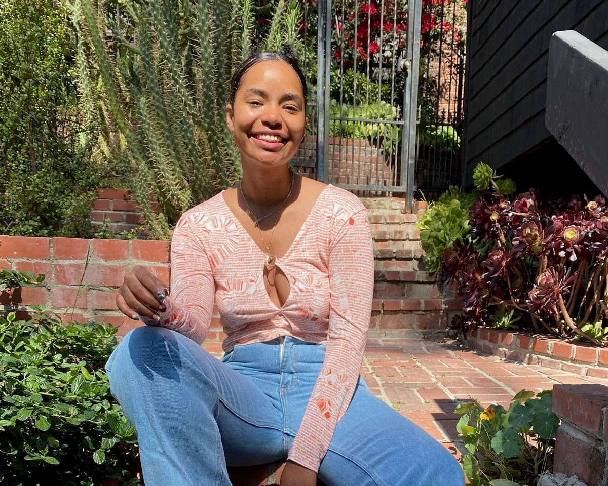 A woman outside smiling sitting in a courtyard surrounded by plants, she's wearing blue jeans and a graphic orange top.