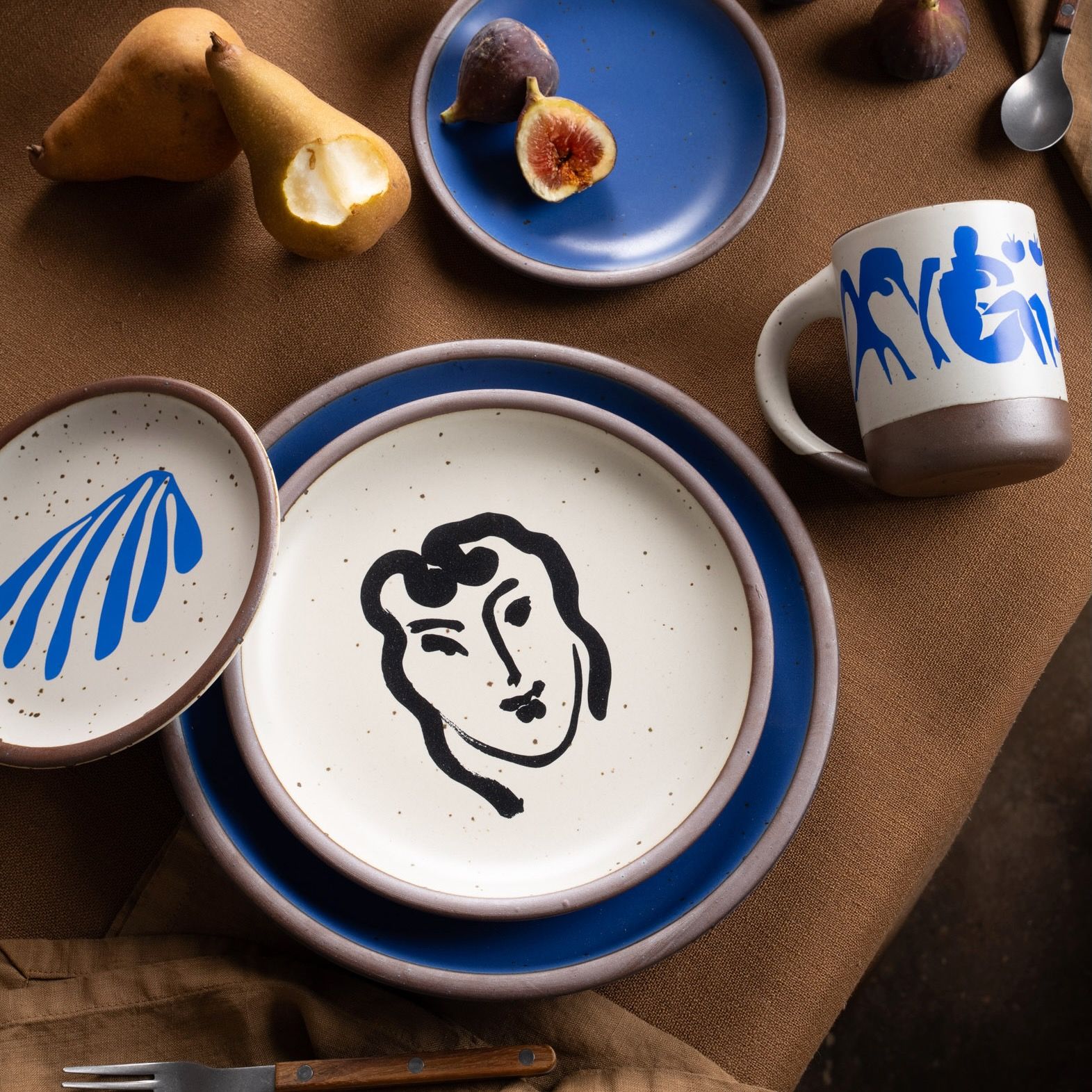 A ceramic plate featuring a decal of the artwork, Patitcha souriante, by Henri Matisse. A minimalist black ink decal of a woman's smiling face in an abstract, expressive style centered on an off-white ceramic plate with an unglazed rim, styled with a blue plate and other dishes on a table with fruit.