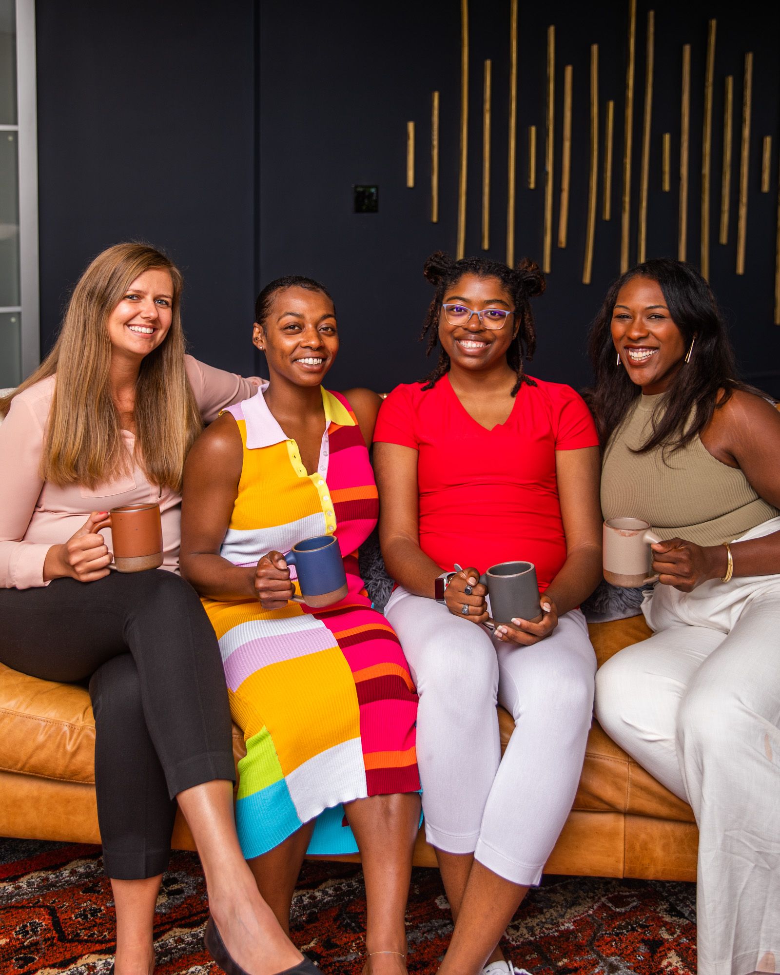 Four people smiling sitting on a light brown leather couch with a black wall background all staring at the camera. Each person is holding a ceramic mug.