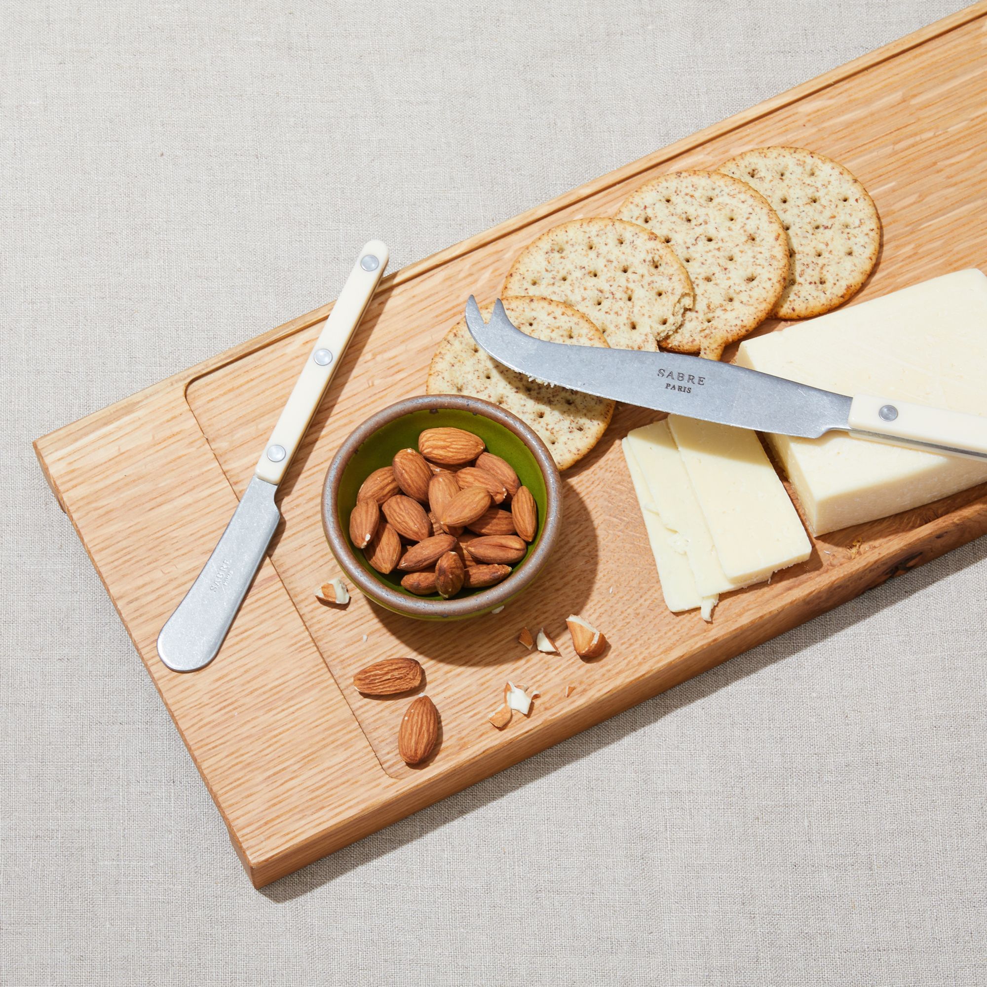 A narrow rectangle of light brown wood with a vertical groove at either end, a small bowl the size of your palm in a fabulous green glaze, two ivory-handled cheese knifes and spreaders, one for hard cheeses, one for soft.