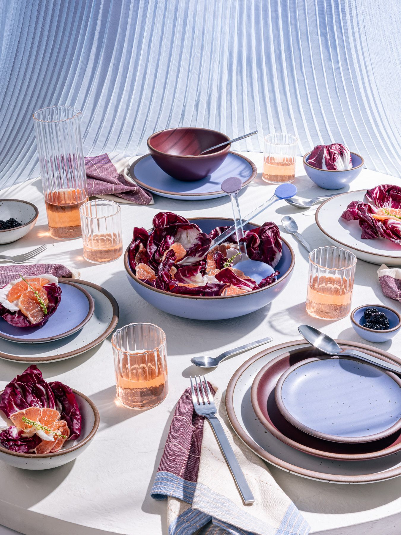 A table filled with ceramic pottery in periwinkle and plum colors, a salad, and glasses filled with orange liquid