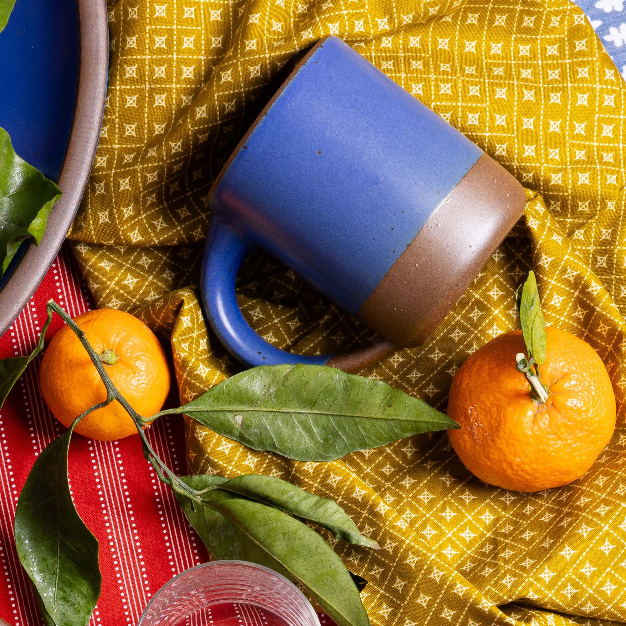 A medium sized ceramic mug with handle in a cobalt blue color featuring iron speckles and unglazed rim and bottom base, against yellow fabric with oranges.