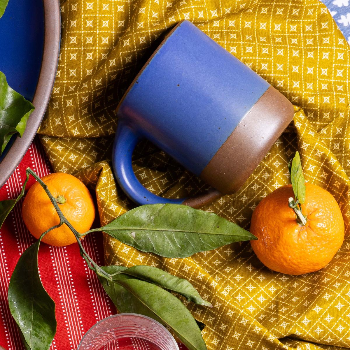 A medium sized ceramic mug with handle in a cobalt blue color featuring iron speckles and unglazed rim and bottom base, against yellow fabric with oranges.