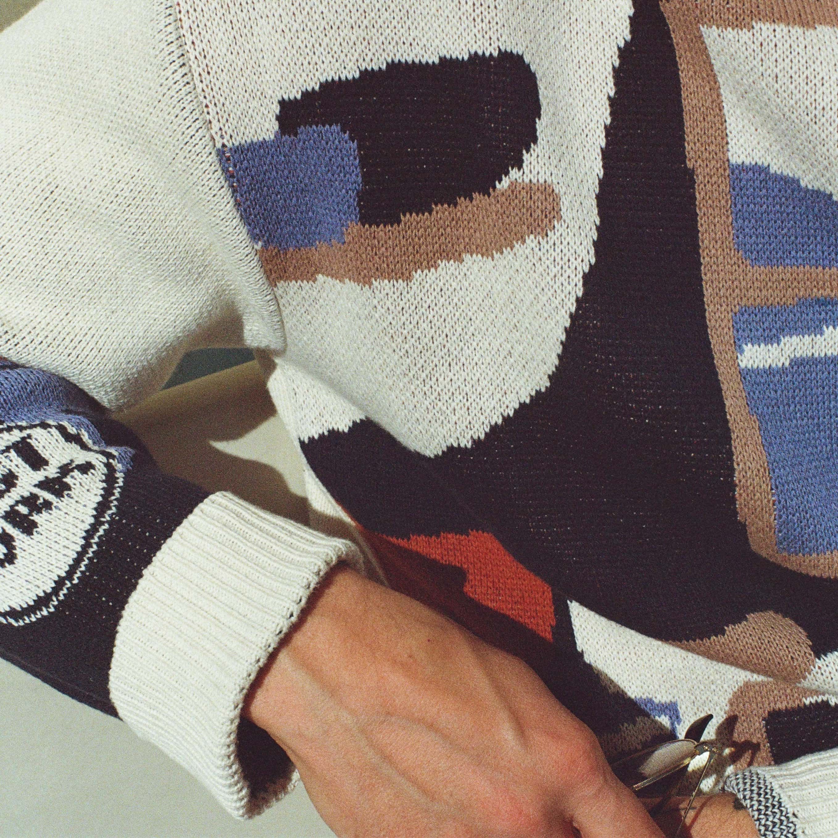 A closeup of a sweater with an illustration of a pottery workshop with a large window, shelves, and table with mugs and bowls. The sweater features cream, navy, terracotta, tan and blue colors.