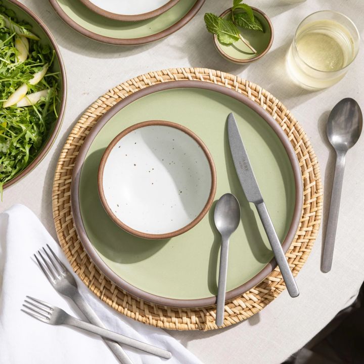 Place setting featuring a rattan charger, a sage green dinner plate, a small cool white bowl, and matte steel flatware.