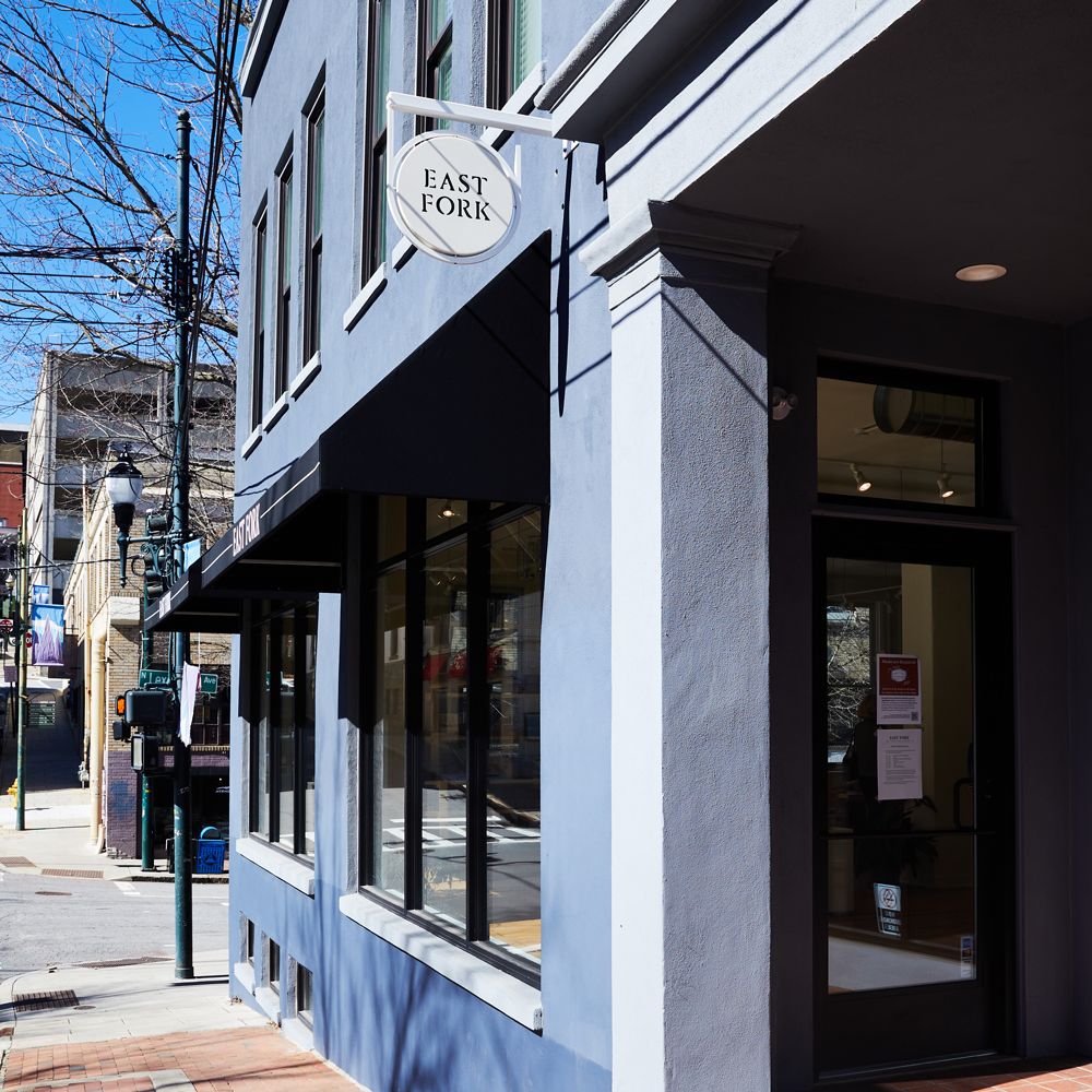 The outside of East Fork Asheville, a blue downtown building with large windows with black awning, and a small white circle sign that reads 'East Fork'.