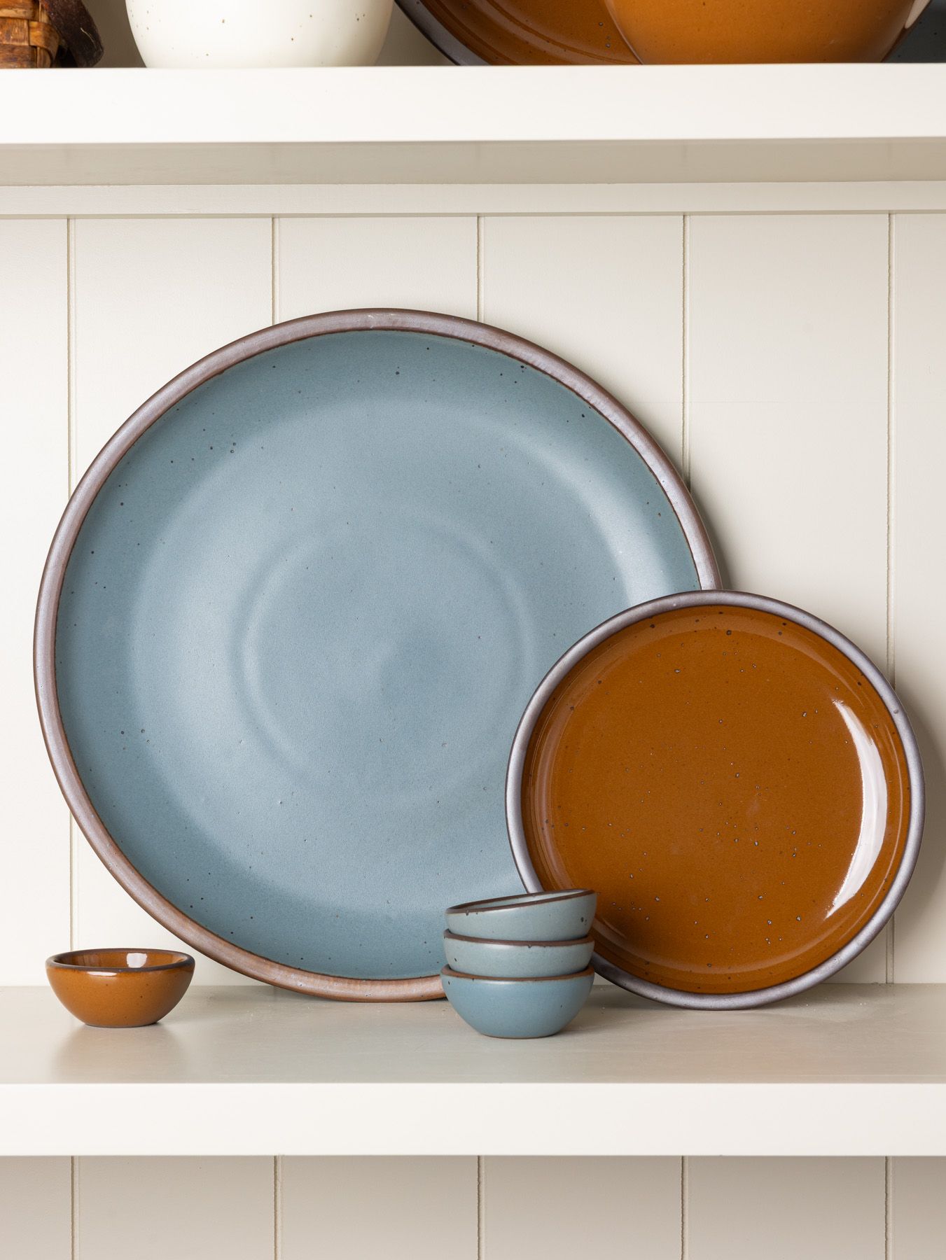 Stacked East Fork pottery in blue-grey and glossy brown colors arranged neatly on a cream shelf against a paneled wall.