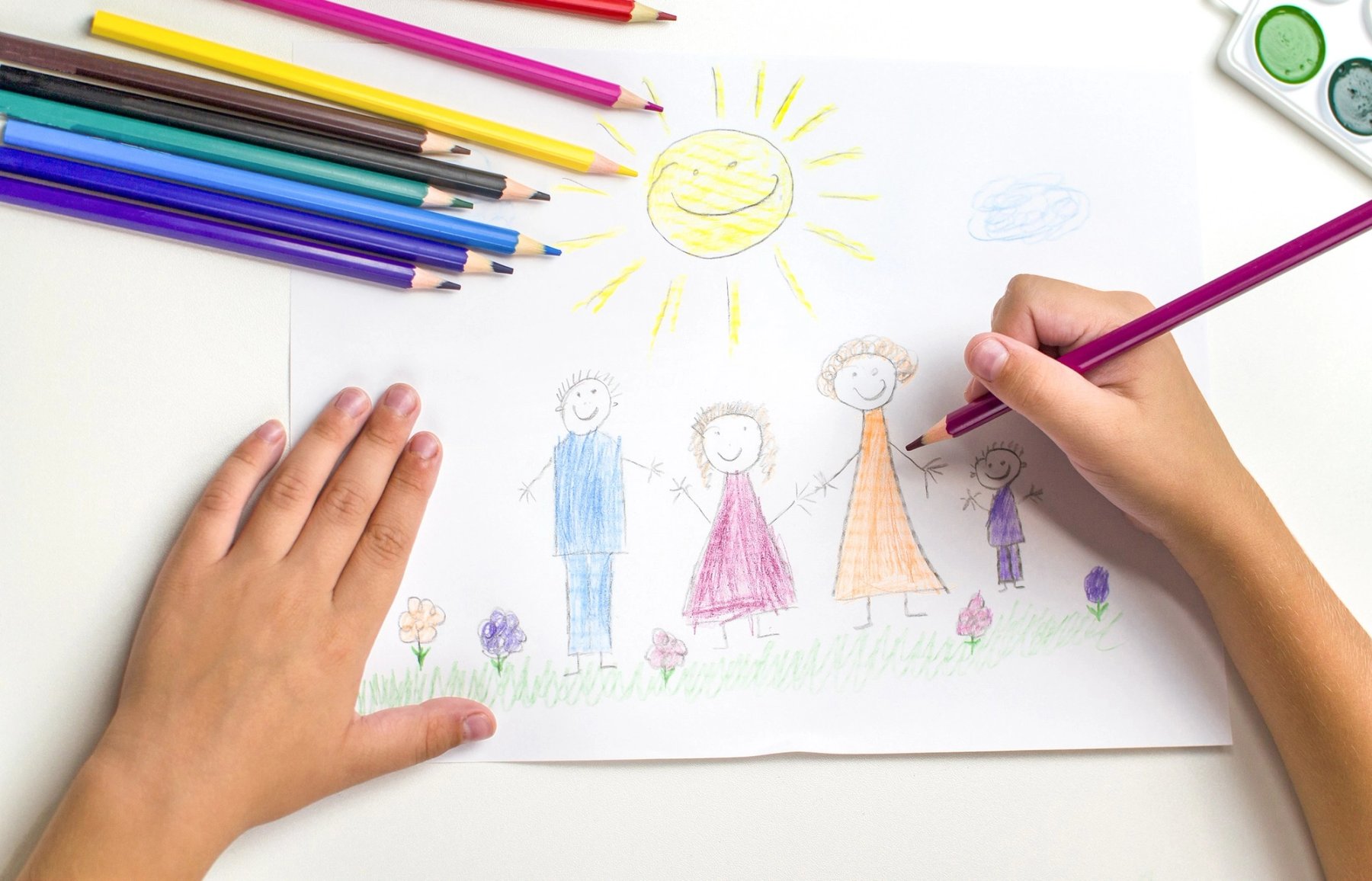 Manos de una niña o un niño dibujando una escena familiar con lápices de colores.