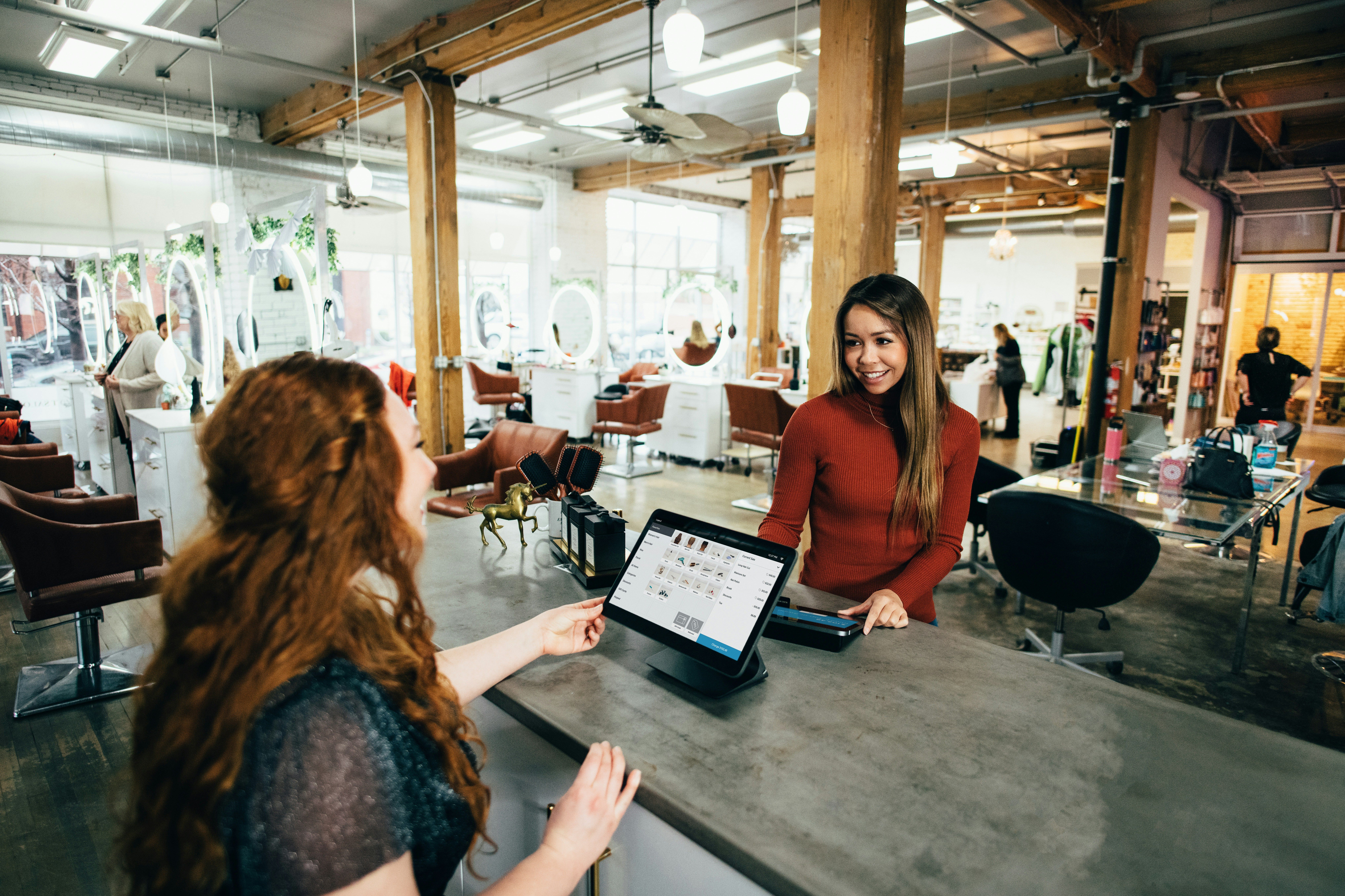 A friendly interaction at a modern hair salon, where a smiling cashier assists a customer at the front desk using a point-of-sale system. The salon has stylish décor, large mirrors, and a lively atmosphere with hairstylists working in the background.