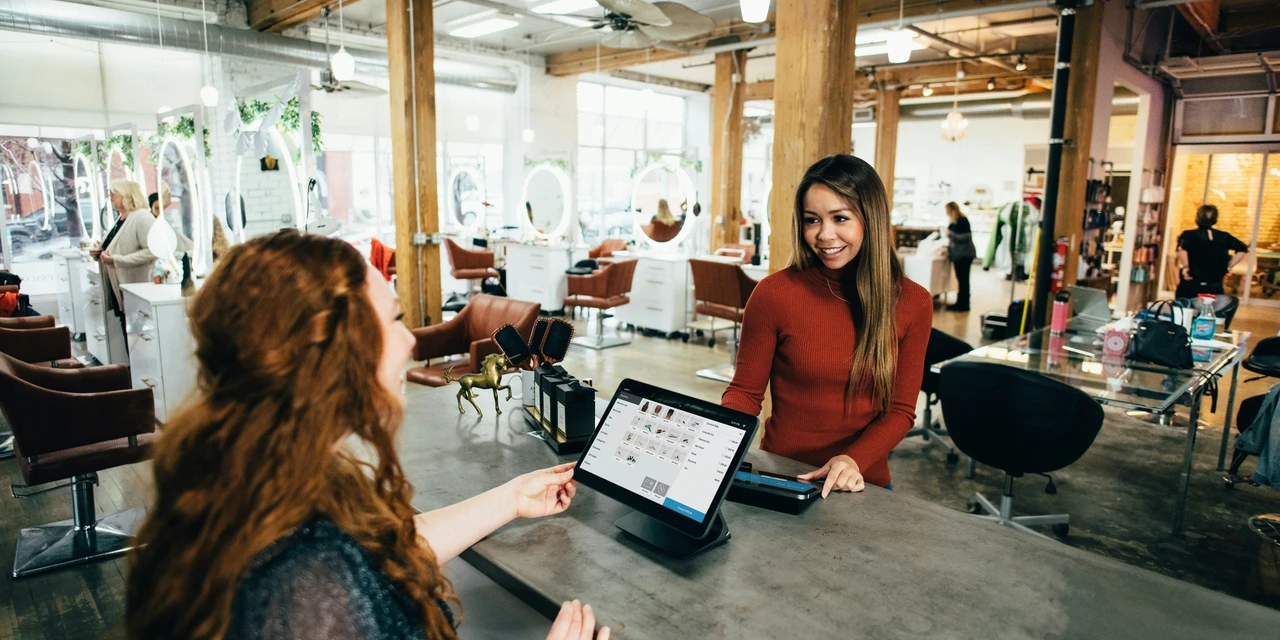 A friendly interaction at a modern hair salon, where a smiling cashier assists a customer at the front desk using a point-of-sale system. The salon has stylish décor, large mirrors, and a lively atmosphere with hairstylists working in the background.