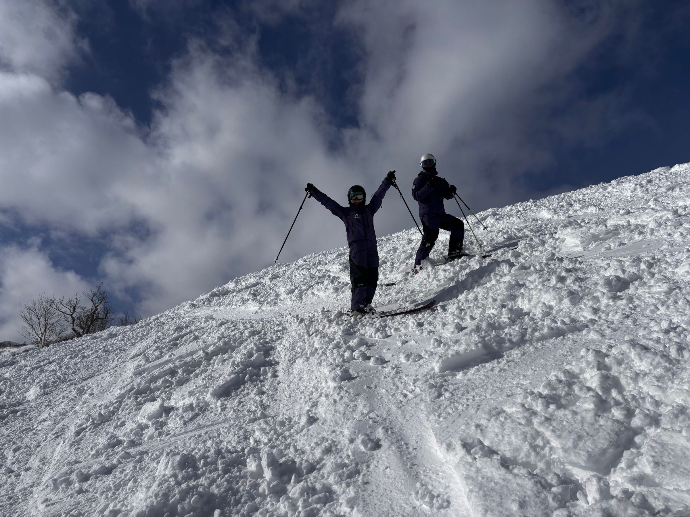 A student throws both arms up on a sunny slope with the coach alongside, blue sky and tracked-out snow behind them