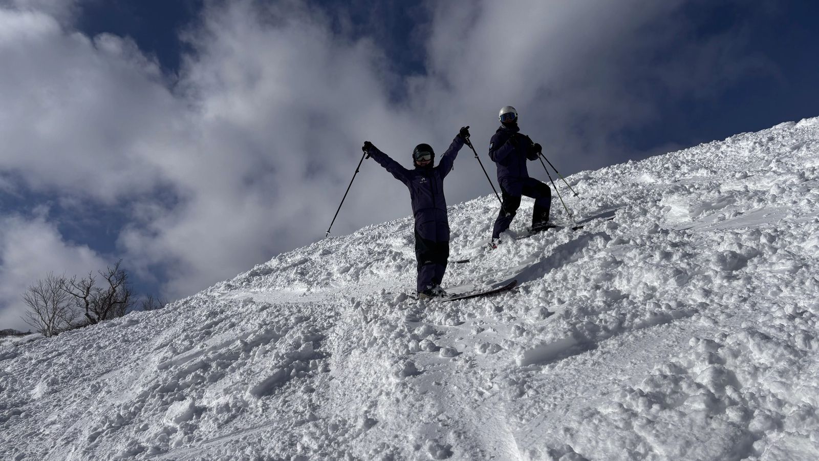 A student throws both arms up on a sunny slope with the coach alongside, blue sky and tracked-out snow behind them