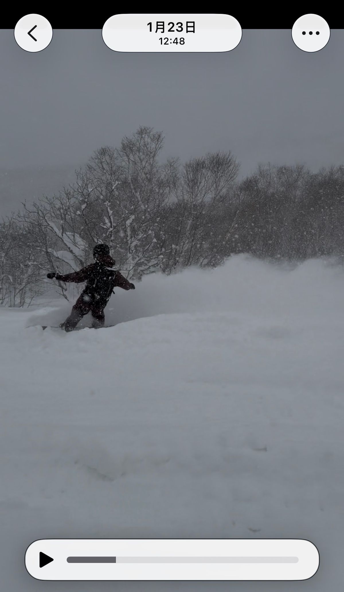A snowboarder turning through deep snow on a snowy day, throwing up a wall of spray. The capture stamp reads 23 January, 12:48