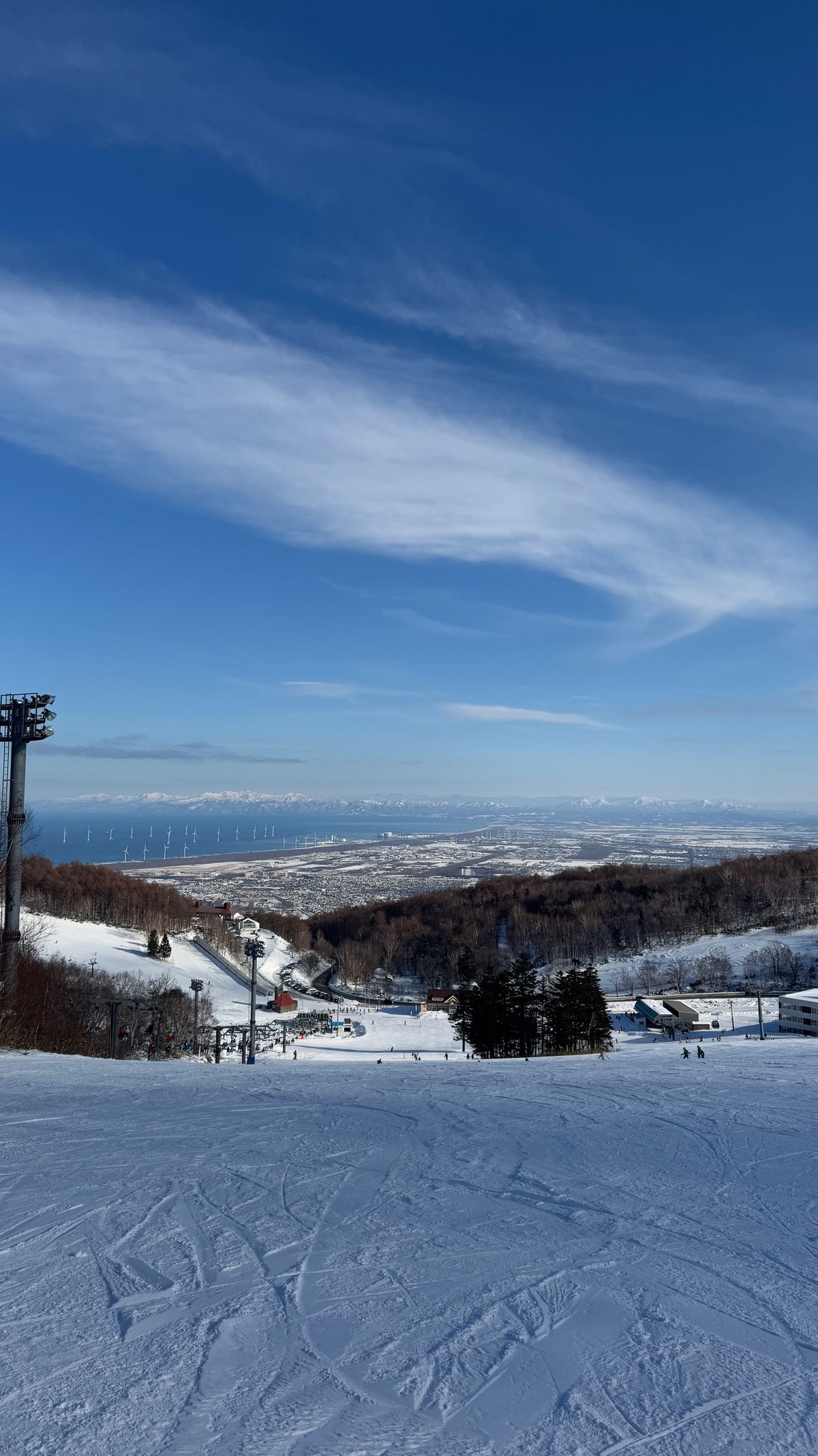 從山頂往下的寬闊雪道,遠處可見石狩灣、風力發電機與山腳的雪場設施