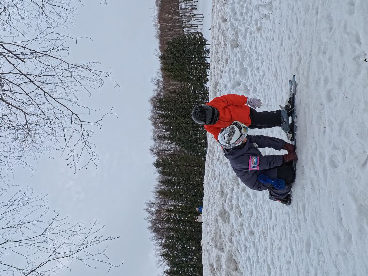 教練蹲下來協助穿橘色雪衣的小朋友確認雪板固定器