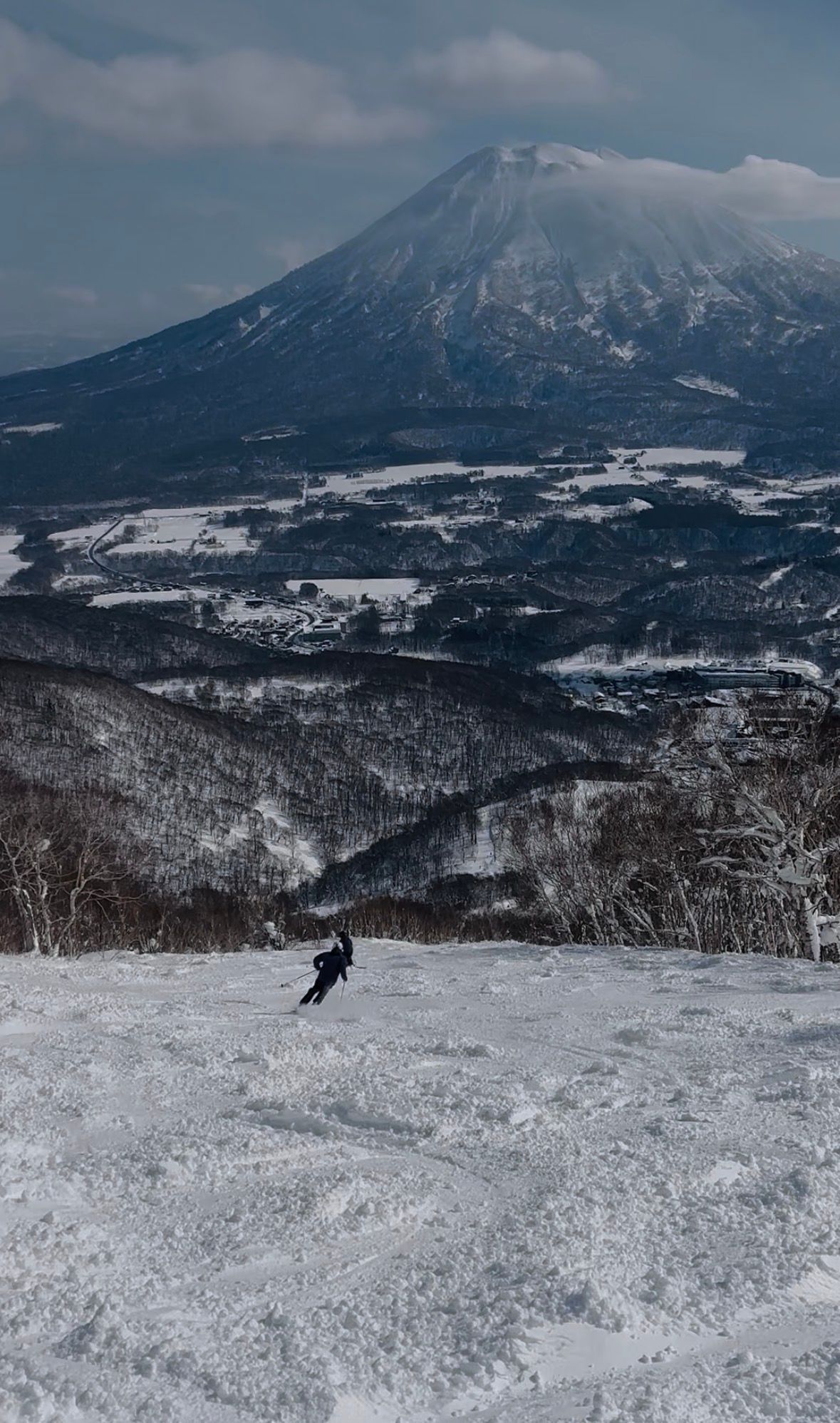 A skier dropping into a wide groomed slope with Mount Yotei filling the horizon behind the valley
