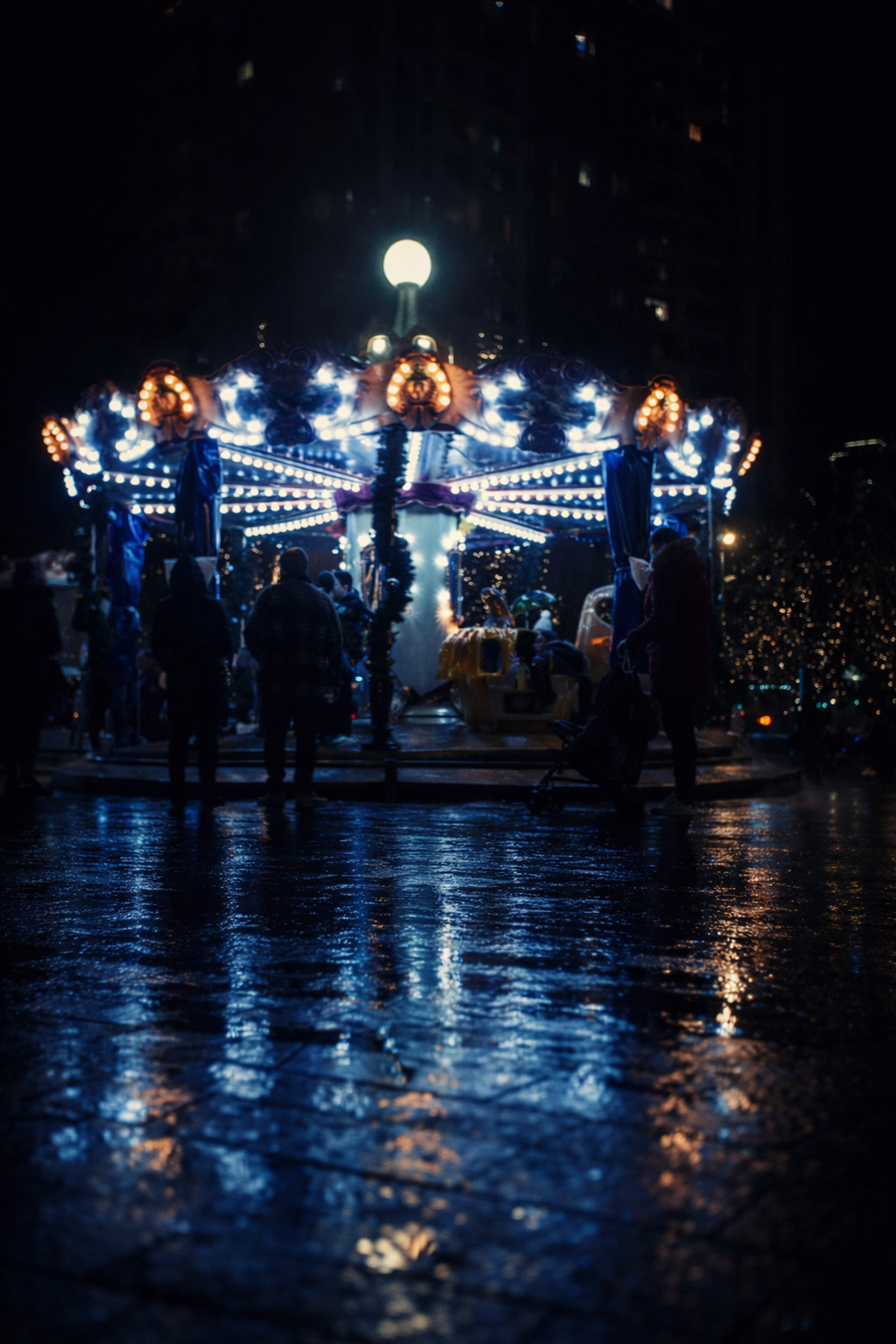 Atmospheric night photograph of a bright carousel glowing with blue and warm lights on a wet city plaza, reflecting brightly across the damp pavement.