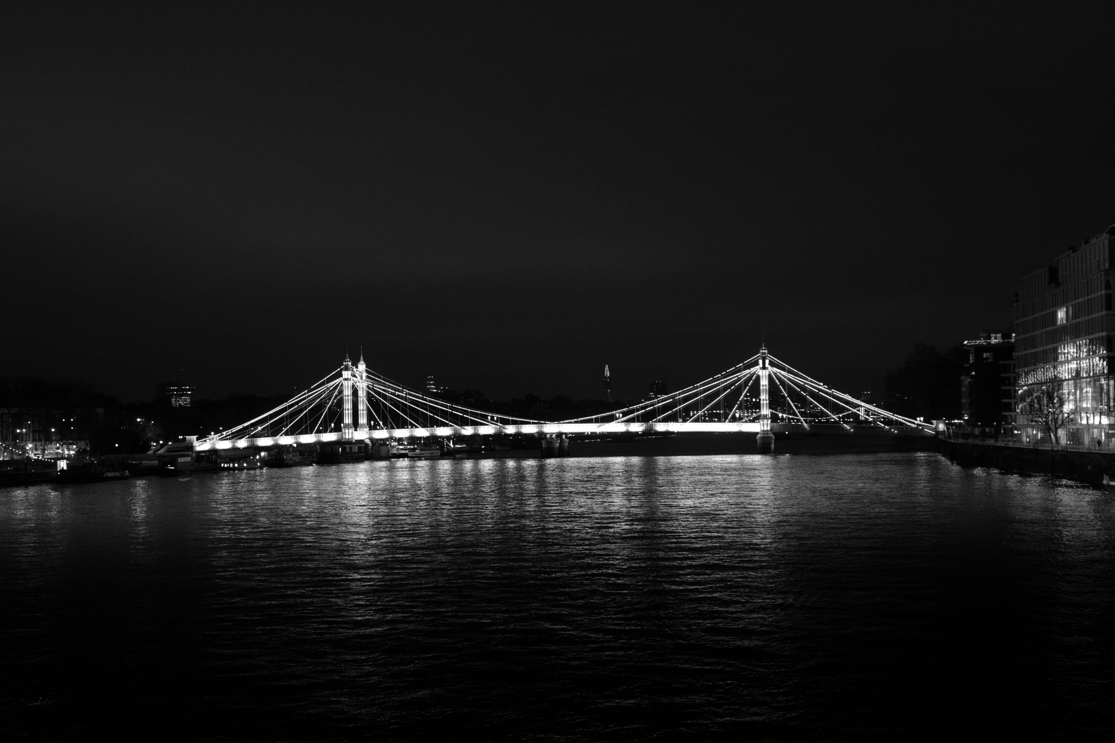 Black and white wide-angle photograph of the illuminated Albert Bridge spanning the River Thames at night, casting glowing reflections on the dark water surface