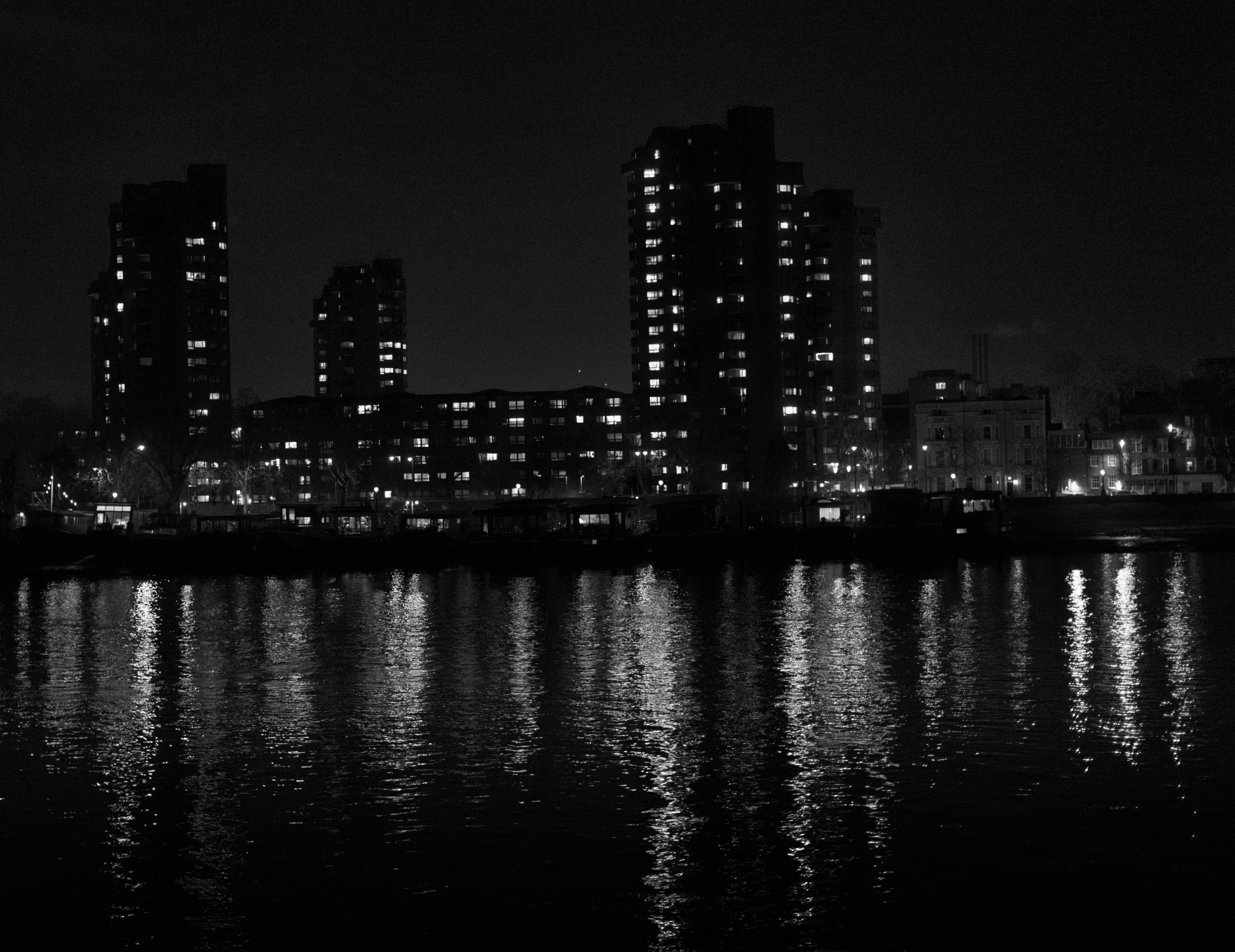 Black and white cityscape photograph of high-rise apartment towers at night across a dark river, with window lights creating long vertical reflections on the water surface.