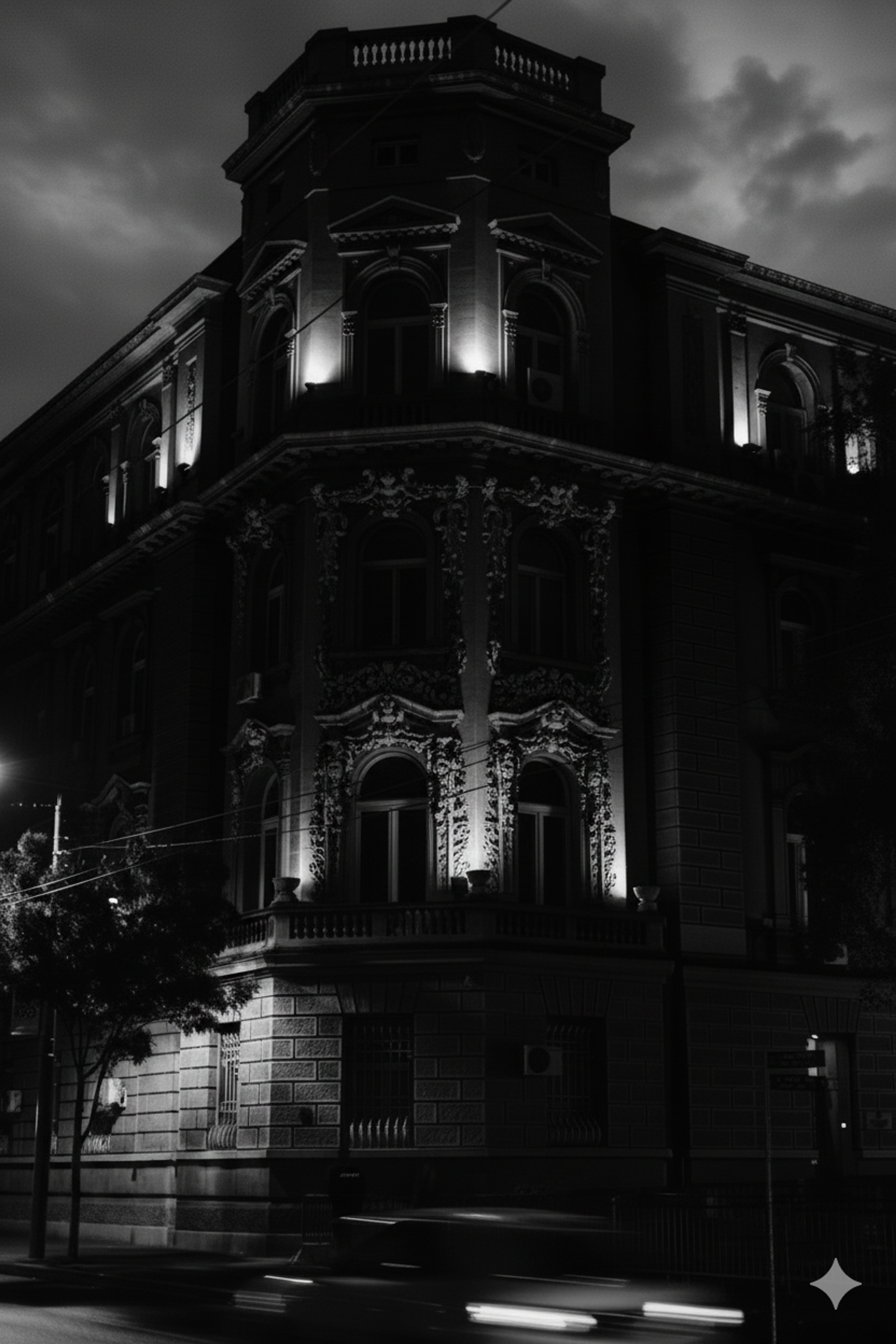 Black and white low-angle photograph of an ornate classical European corner building illuminated by upward-facing night lights against a dark sky. 