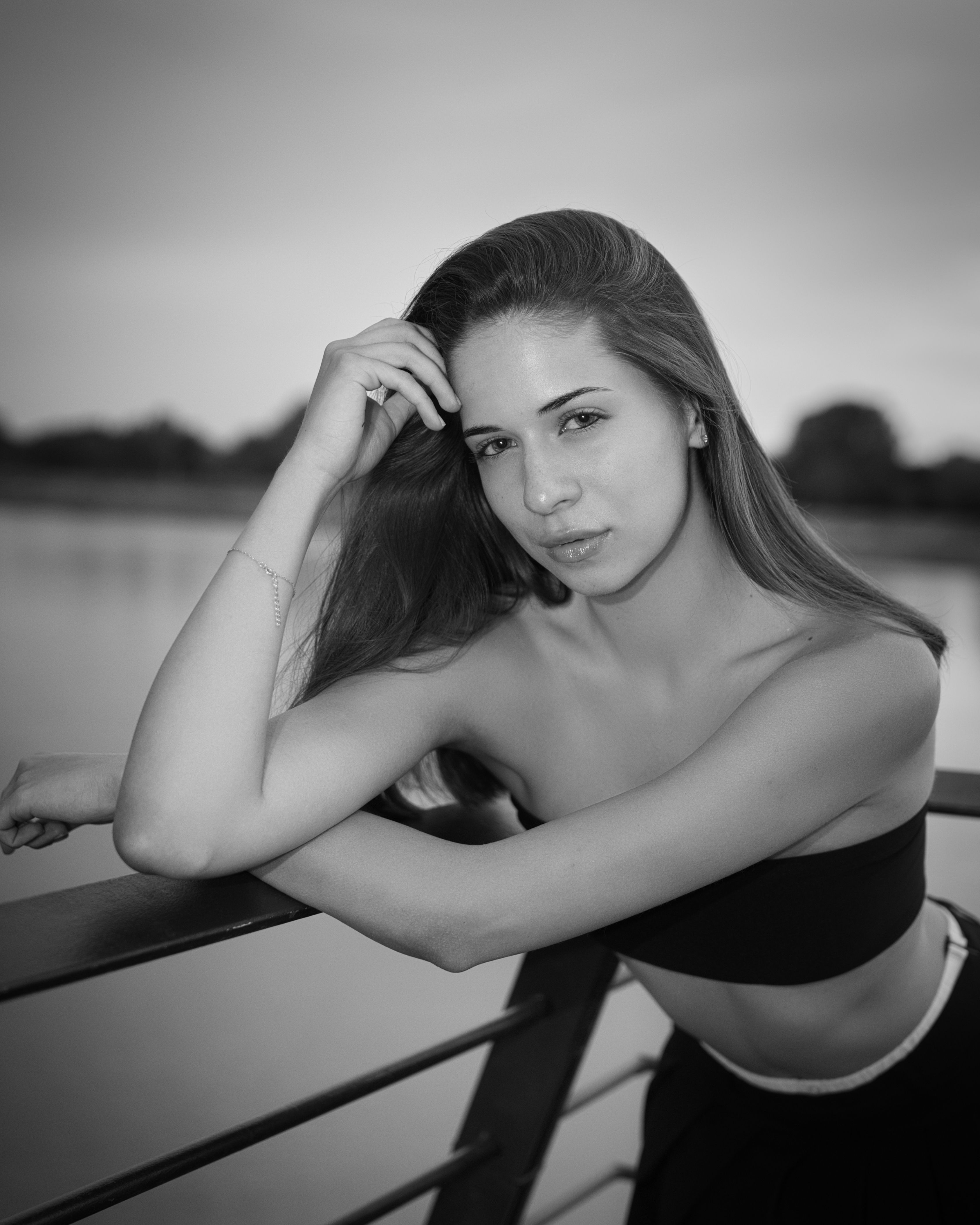 Black-and-white outdoor portrait of a young woman leaning on a railing by a body of water, gently running her fingers through her hair.