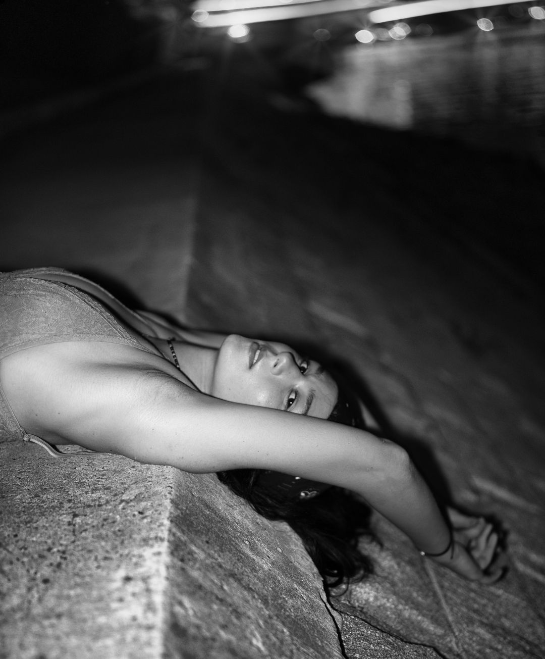 Dramatic monochrome photograph of a woman reclining against an outdoor concrete structure at night with moody lighting.