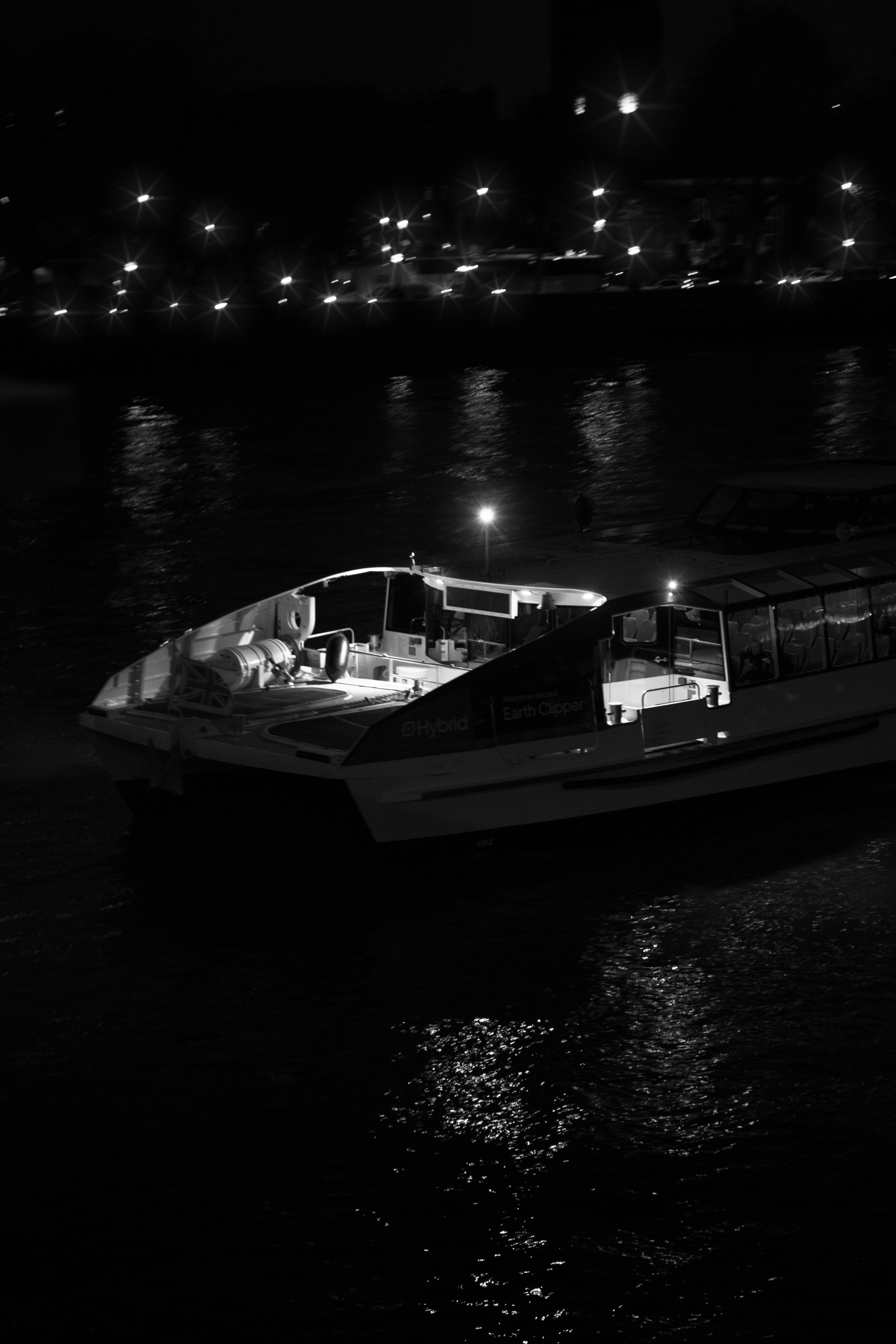 Black and white vertical photograph of an illuminated passenger ferry cruising down a dark river at night with distant shore lights reflecting on the water.